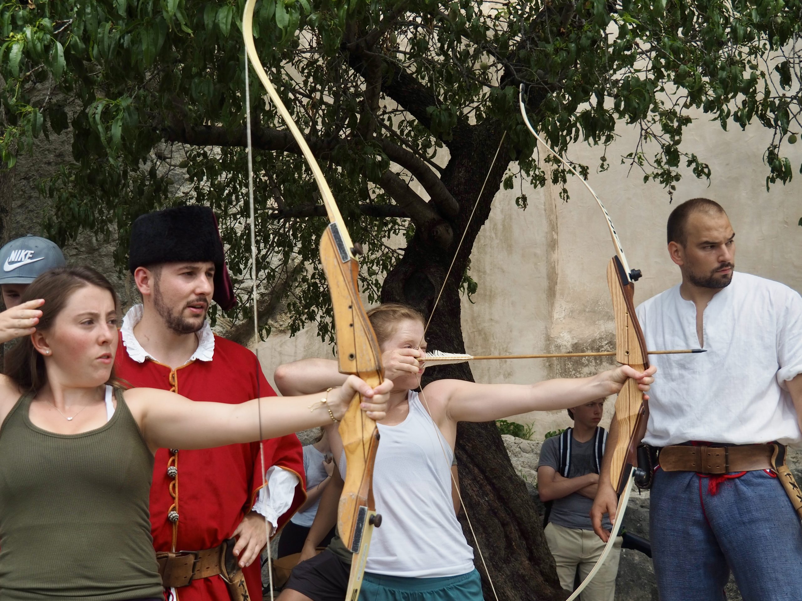 Two students draw traditional bows under a tree while guides in period costume watch and coach them.
