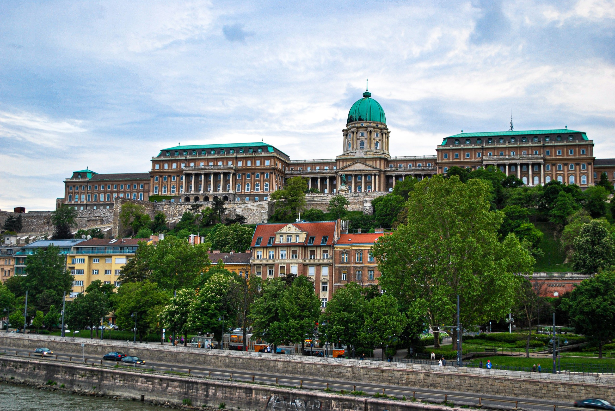 Buda Castle's green dome and long colonnaded wings rise above trees and river-front houses on a summer afternoon.
