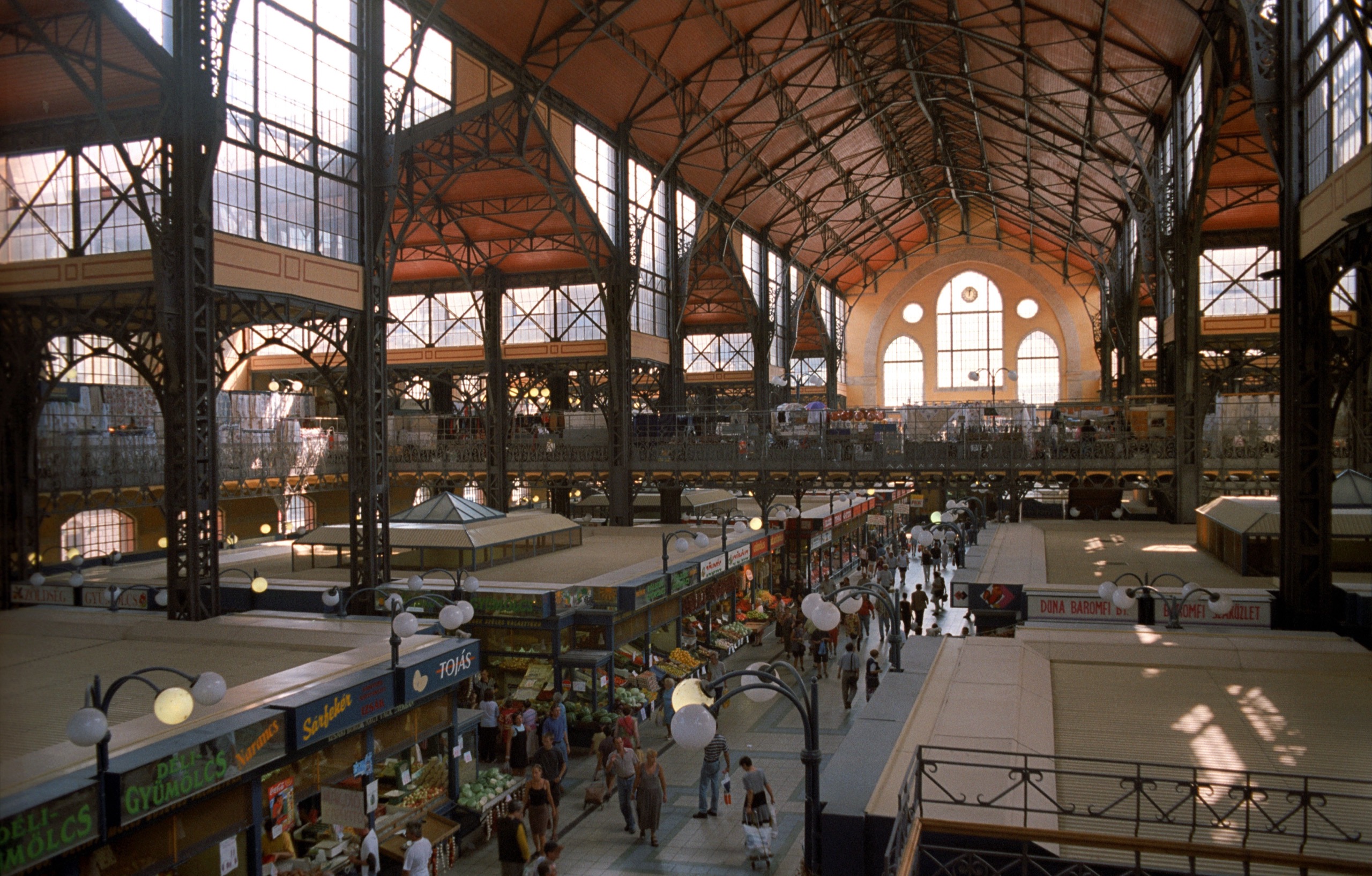 Inside the Great Market Hall, iron roof trusses above rows of produce stalls and shoppers, sunlight falling through the end window.