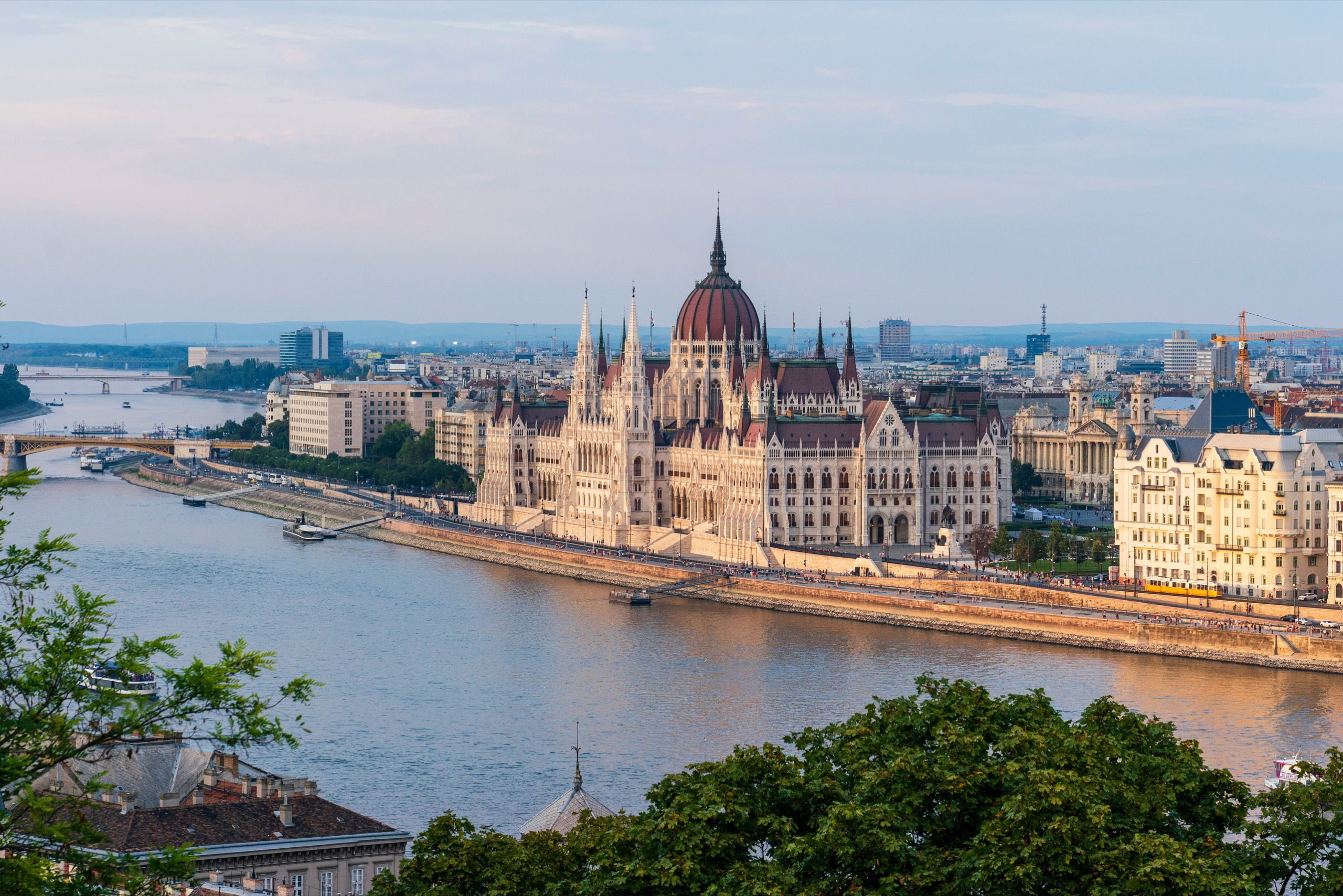 The Hungarian Parliament in Budapest, Hungary, seen across the Danube at dusk, its red dome and white spires above the river and a bridge beyond.