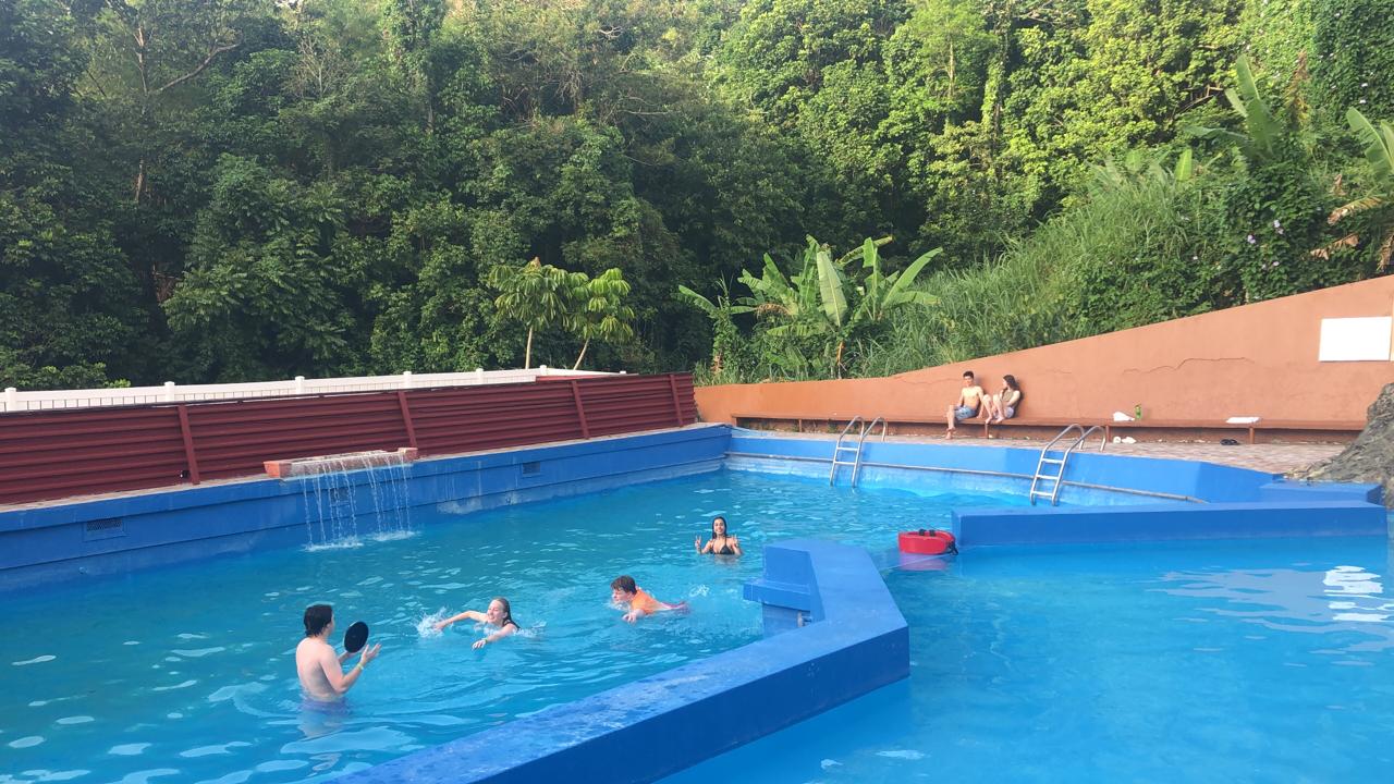 Students swim and relax in a blue pool surrounded by lush forest