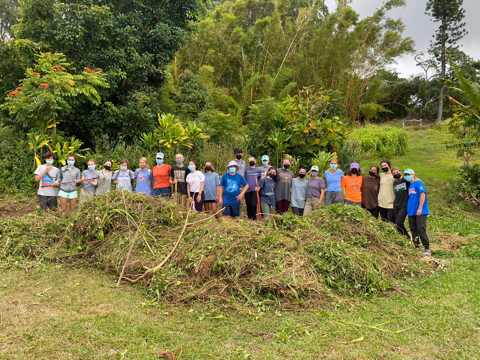 Teen travelers in masks stand behind a large pile of cleared plant debris on a farm