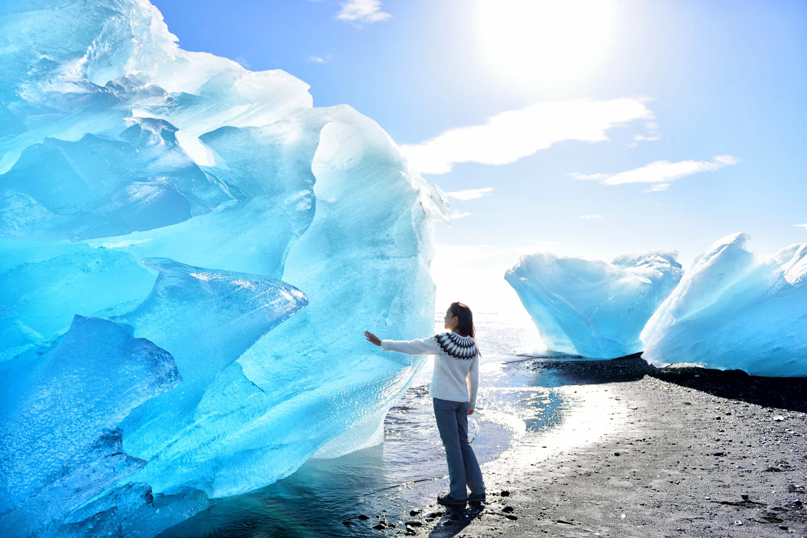 A traveler in a patterned wool sweater reaches out to touch a glowing blue iceberg on a black sand beach