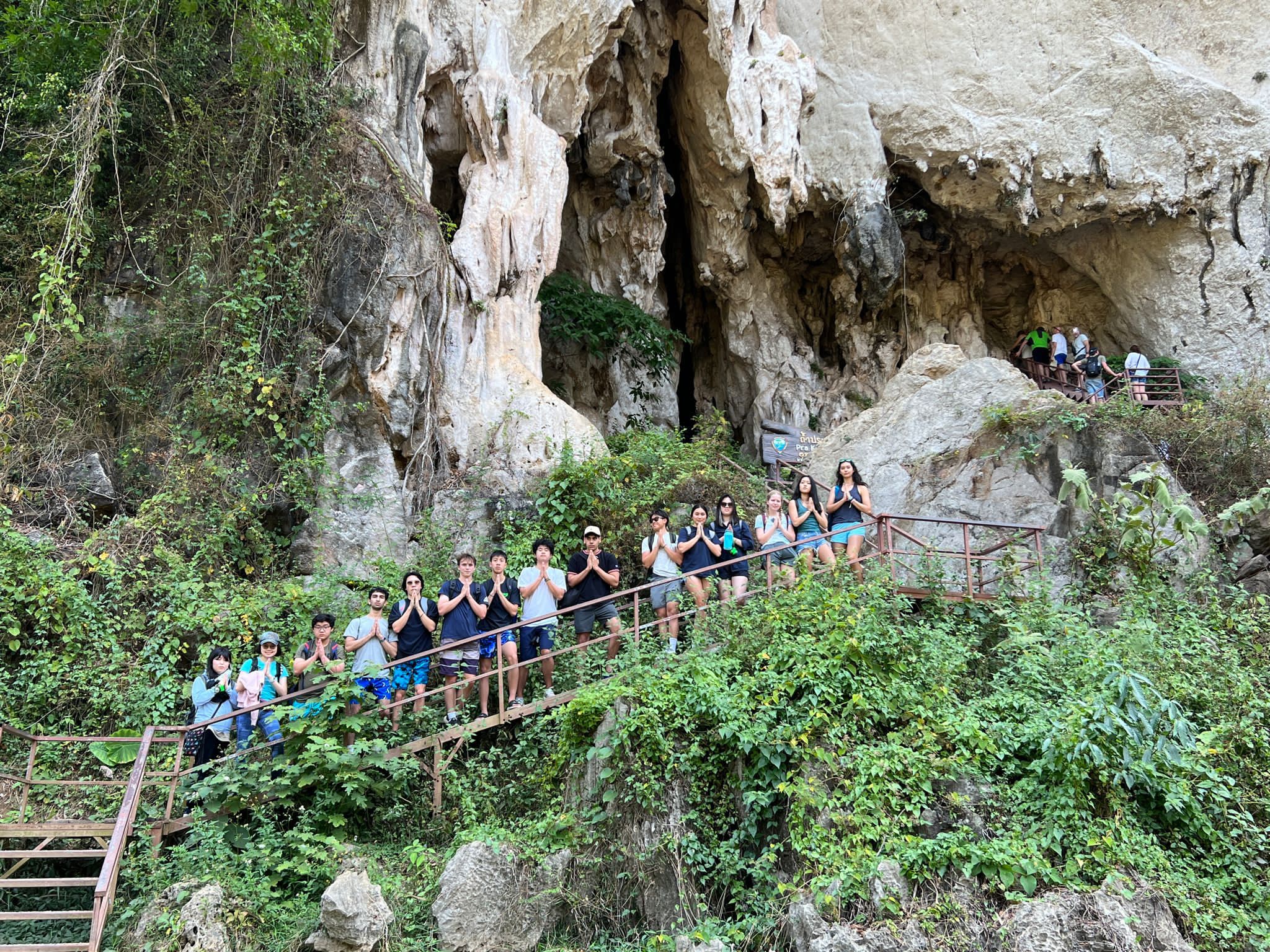 Students pose with hands together on a wooden walkway beneath a limestone cave entrance in Thailand