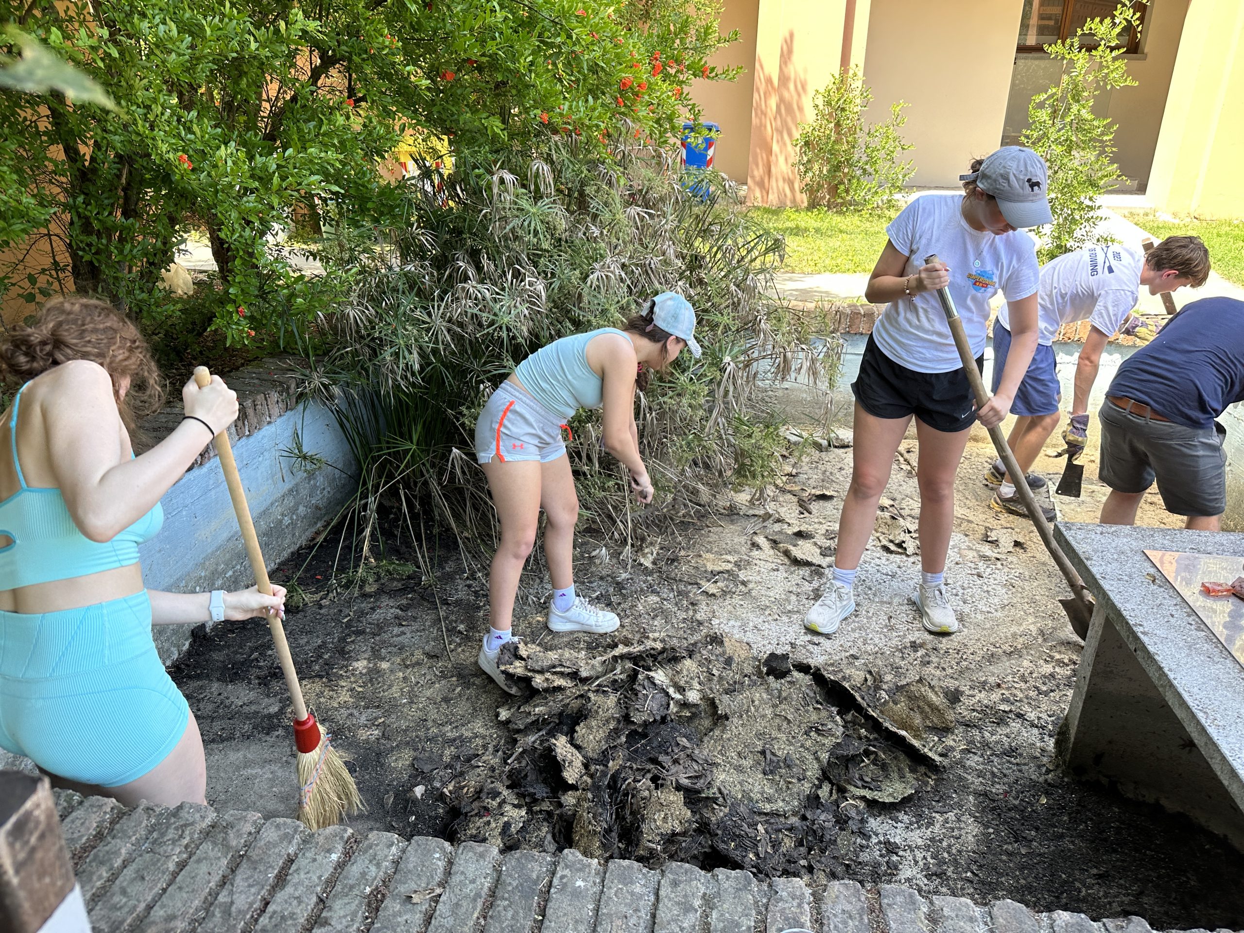 A school trip service project has students clearing debris and digging with shovels and a broom