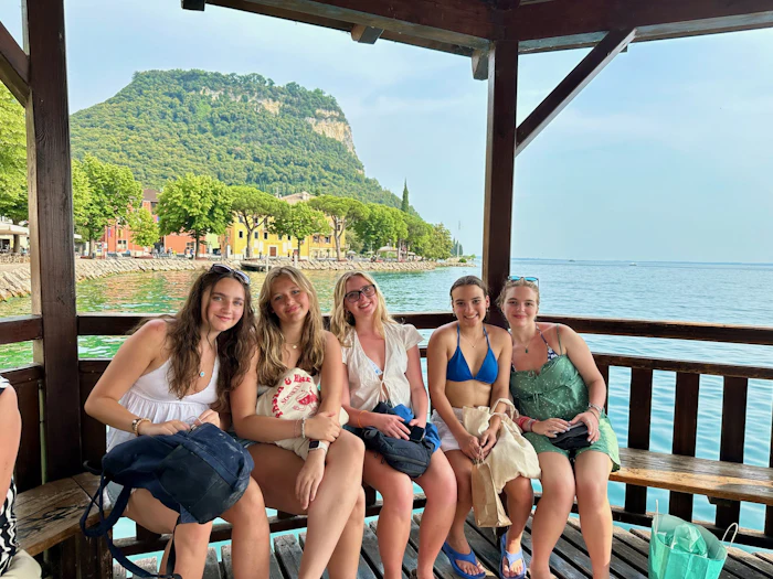 Students sit along the rail of an open lakeside pavilion, with steep wooded slopes and a cluster of pale houses on the far shore.