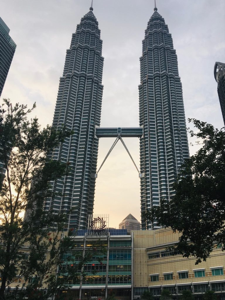 The high Petronas Towers standing behind green trees in Kuala Lumpur. The picture captures the sunset happening in the capital city of Malaysia.