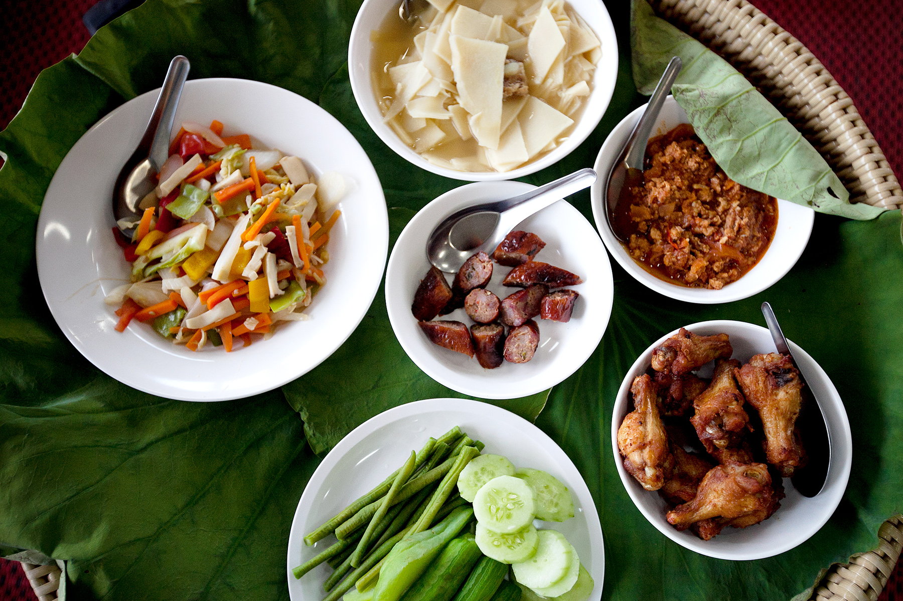 Bowls of Thai dishes including salad, chicken wings, sausage, and dipping sauces arranged on a banana leaf