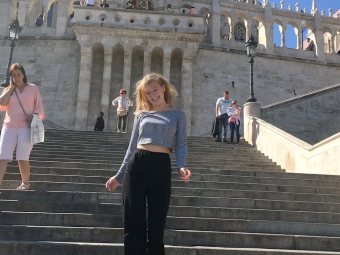 A student walks down the wide stone steps of the Fisherman's Bastion, its white turrets and arches behind her in bright sun.