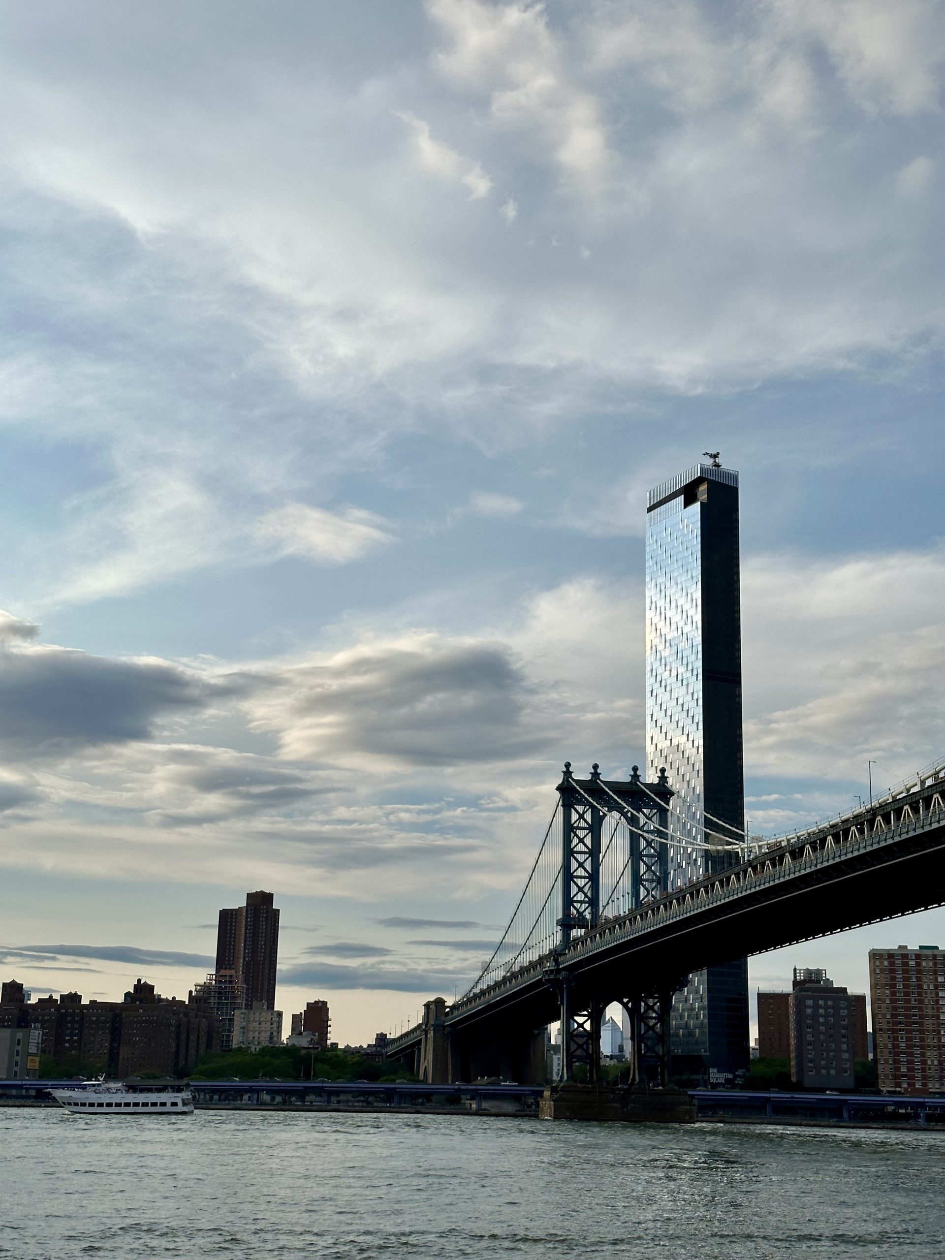 A school trip to New York City pauses as Manhattan Bridge spans the East River beneath a cloudy sky with a skyscraper rising behind it