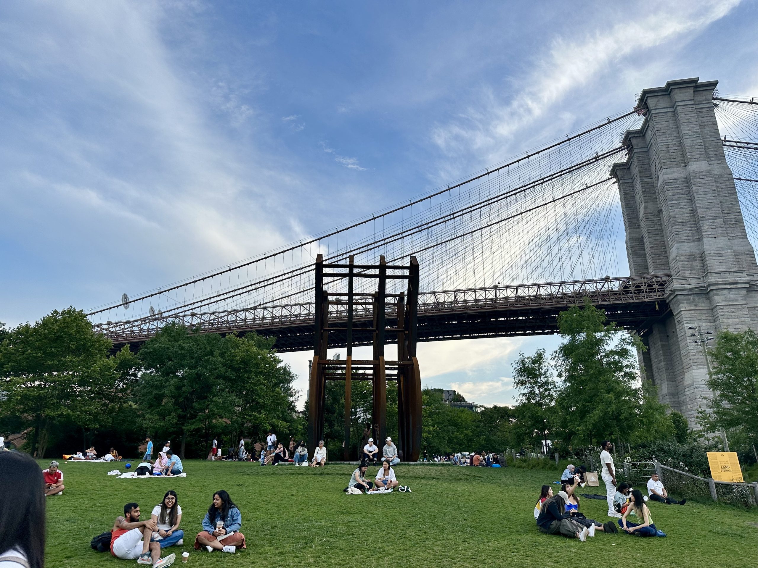 People relaxing on the grass at Brooklyn Bridge Park with the bridge overhead.