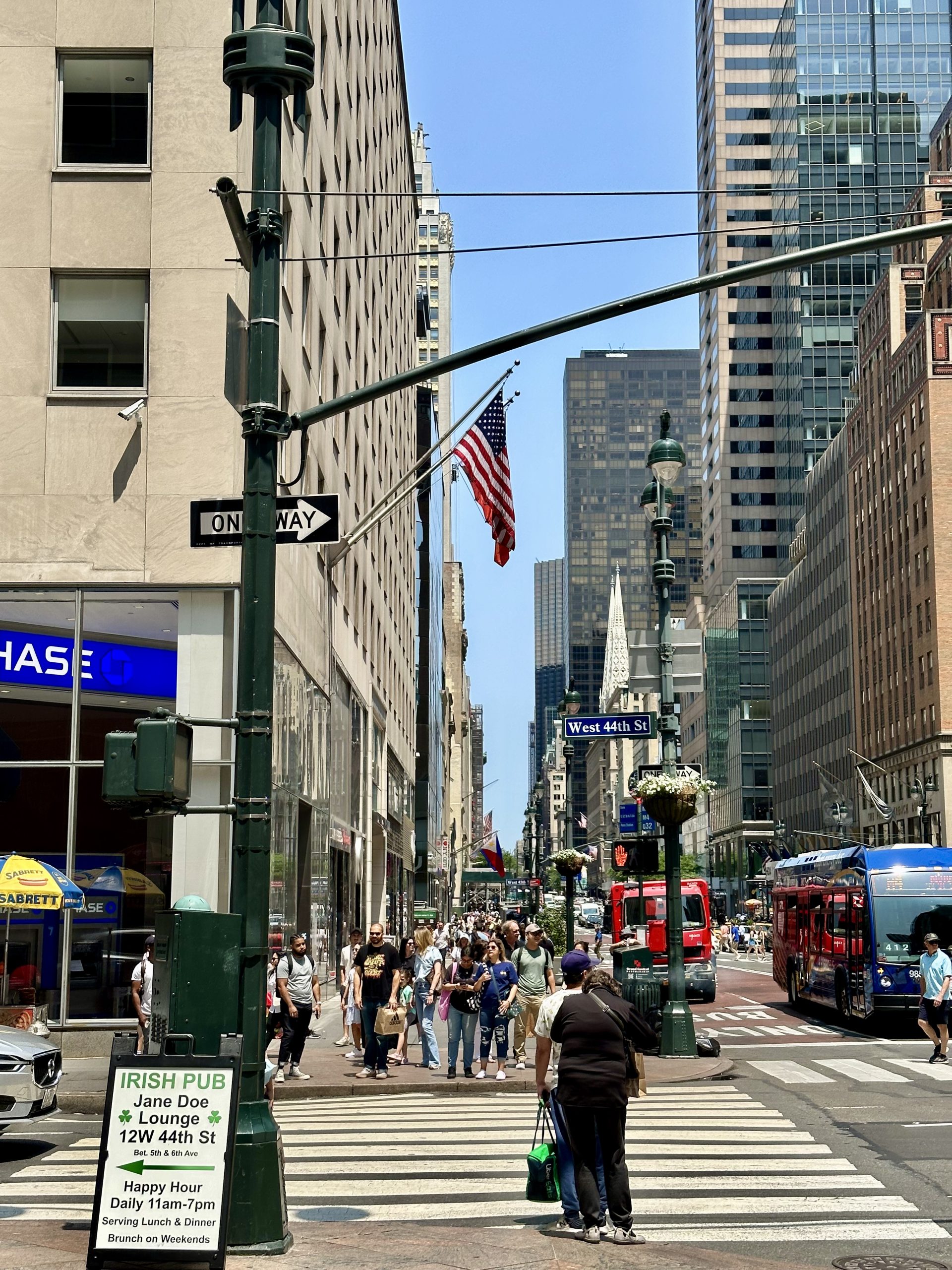Crowded street scene near West 44th St with the American flag and surrounding high-rise buildings.