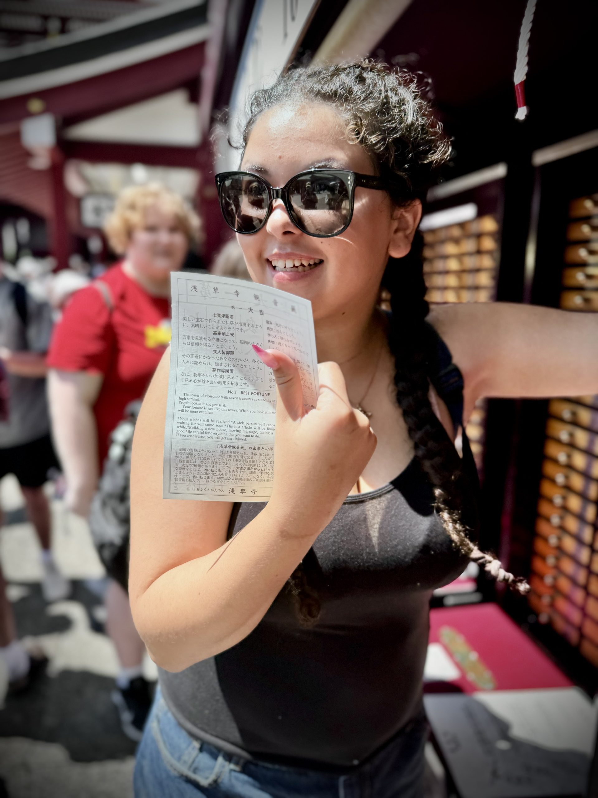 A smiling woman wearing sunglasses holds up a fortune slip at a bustling Japanese temple. fun facts about tokyo