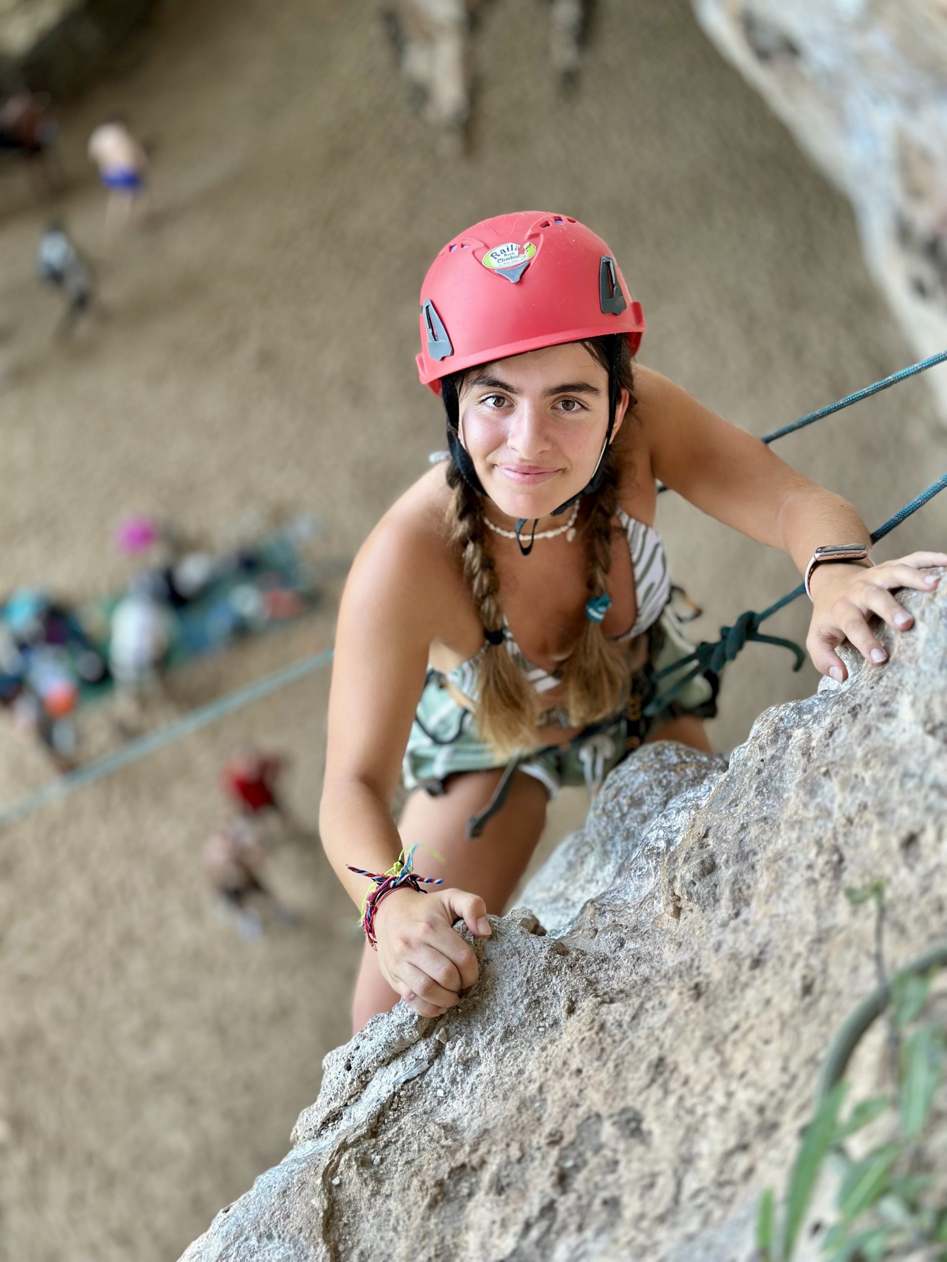 A student in a red helmet climbs confidently up a vertical rock wall in Thailand, with the sandy beach and other climbers visible far below.