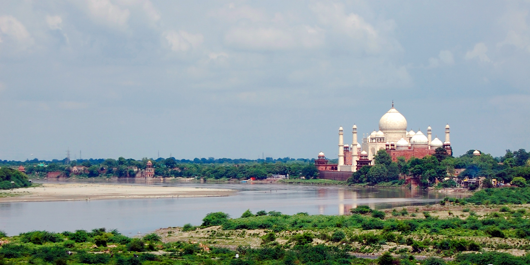 The Taj Mahal rises above a riverbank landscape under a wide sky