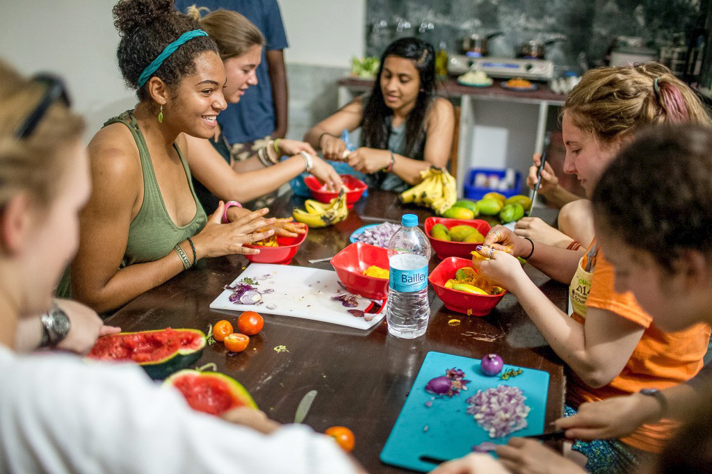 On a school trip, students gather around a table cutting fruit and vegetables into red bowls