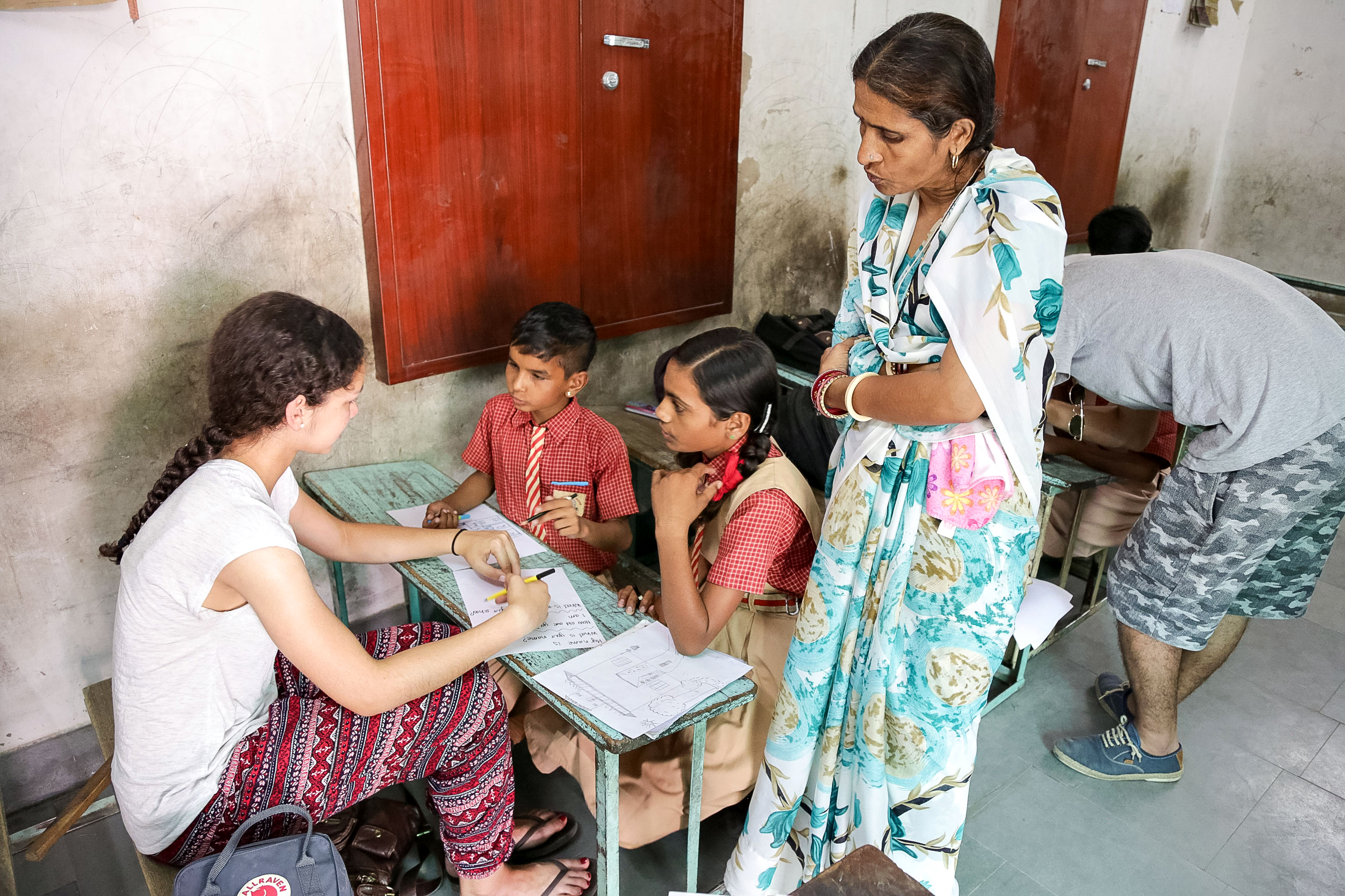 Students help a schoolchild with schoolwork at a desk while a teacher looks on