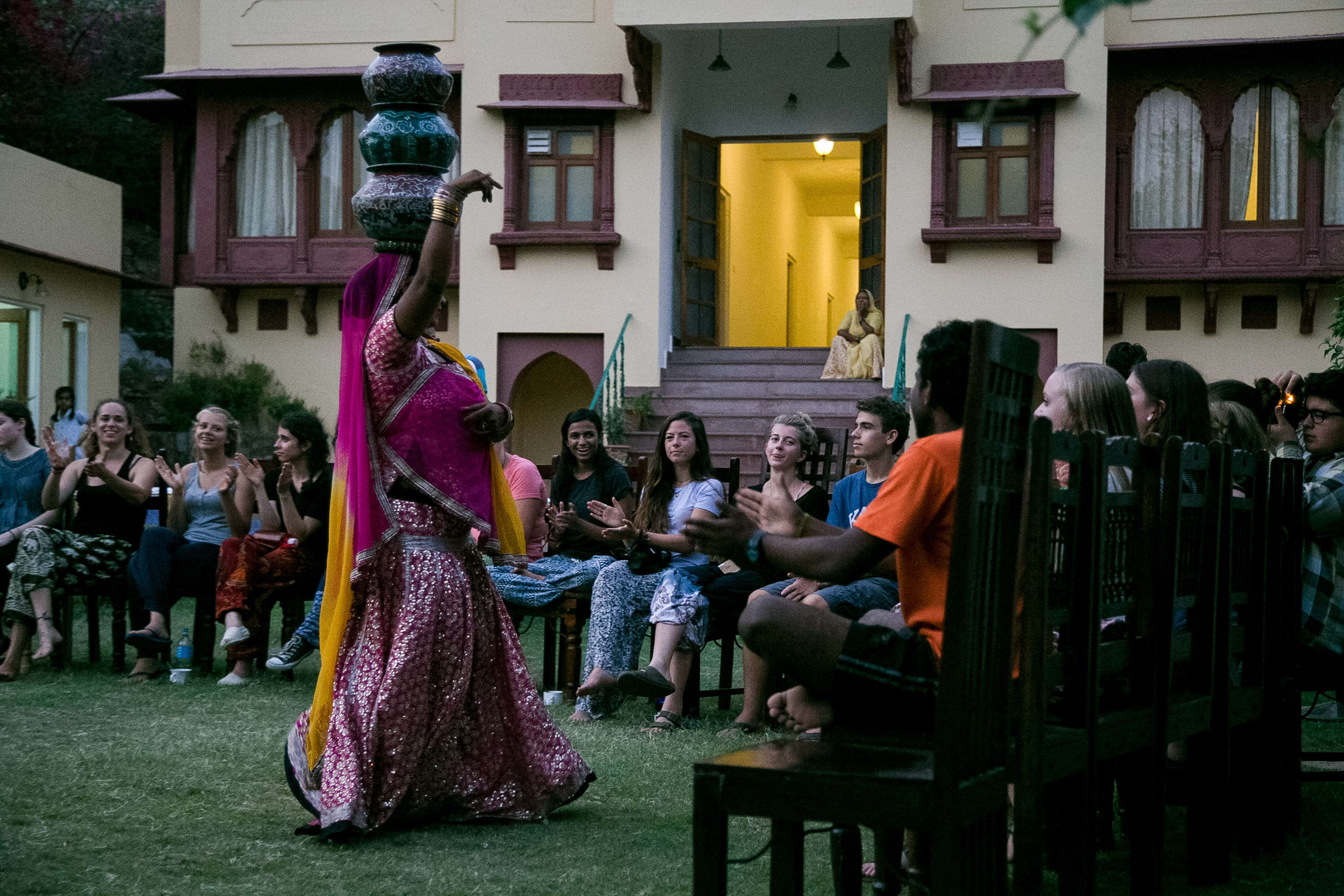 A traditional dancer balances stacked pots on her head while students watch and clap outside