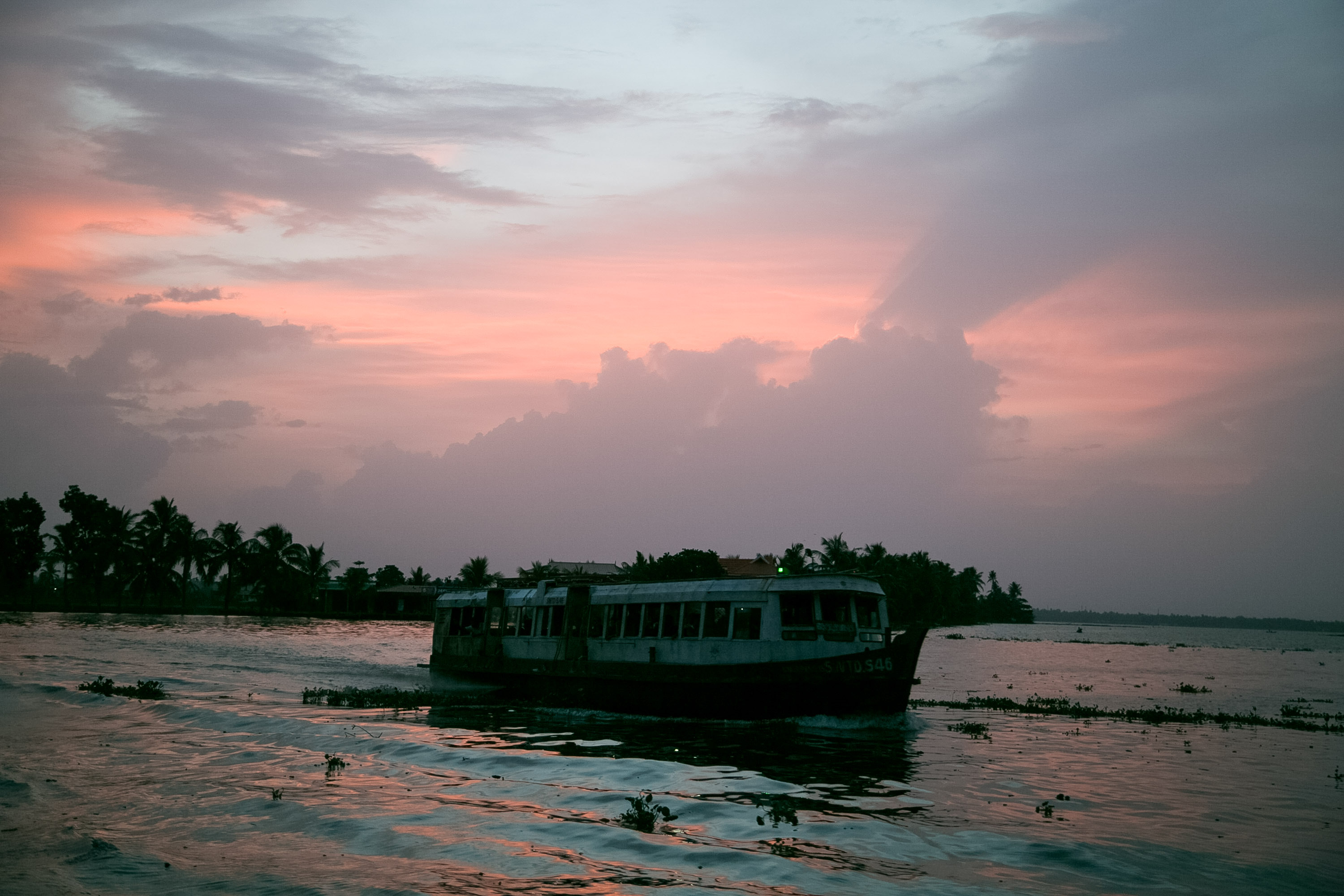A houseboat drifts on calm water in India under a pink and gray sunset sky