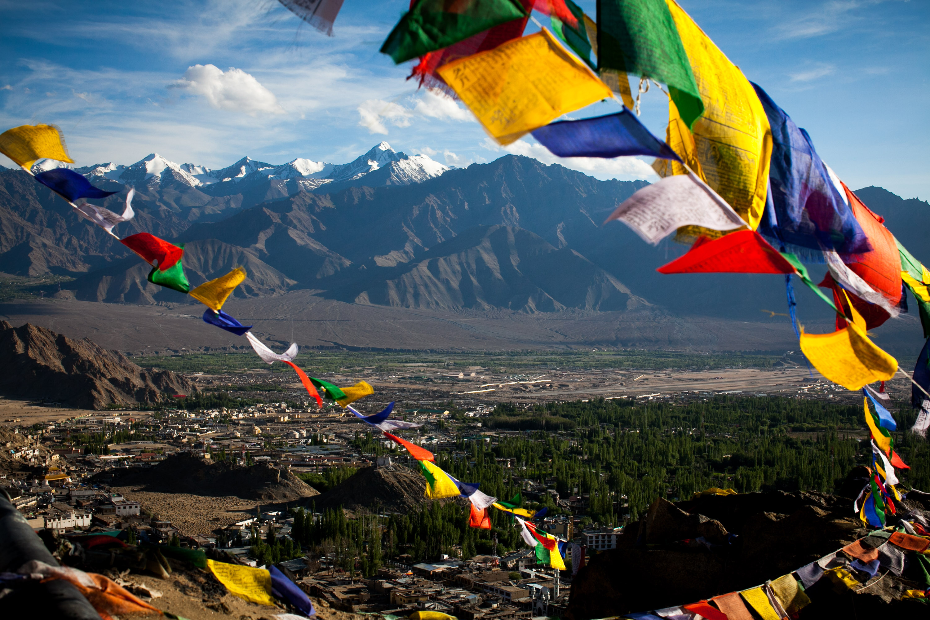 Prayer flags strung above the Indus valley, with the town on the green valley floor and a snow-topped range behind.