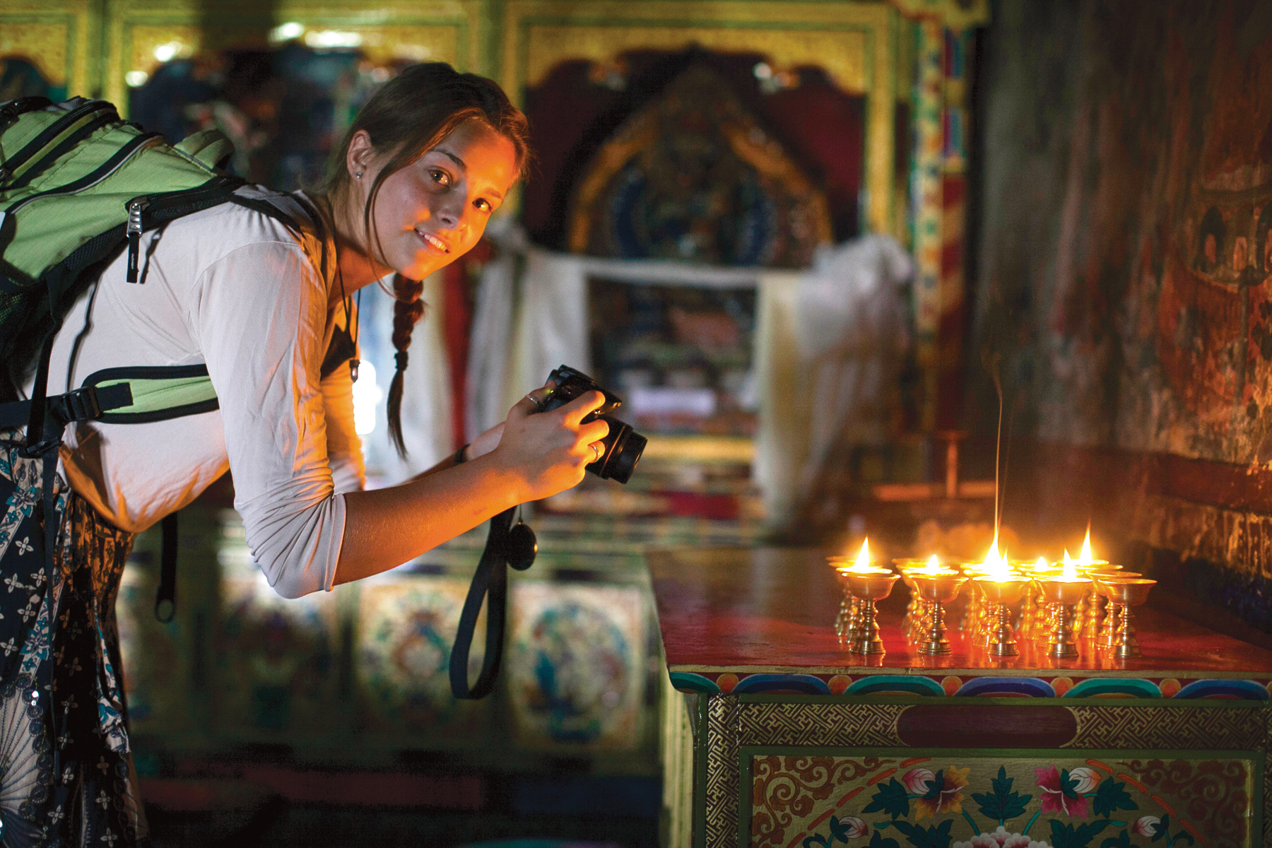 A student with a camera composes a shot beside a row of lit butter lamps in a painted monastery interior.