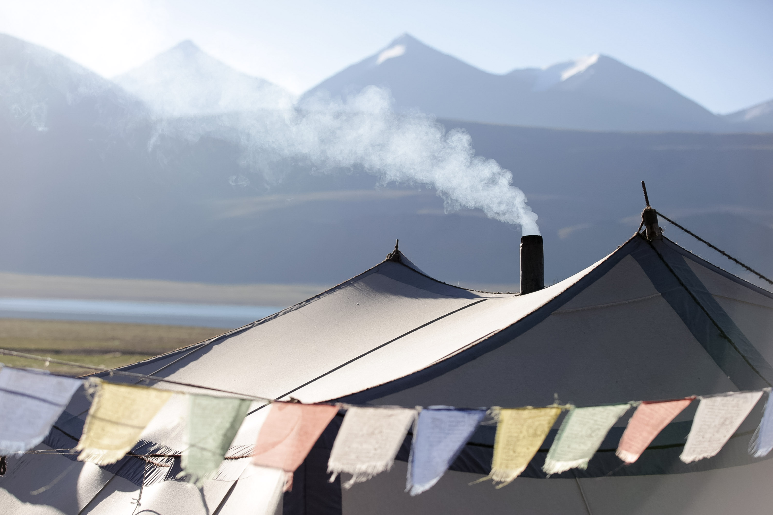 Smoke rises from a tent chimney with prayer flags strung below and Himalayan peaks behind