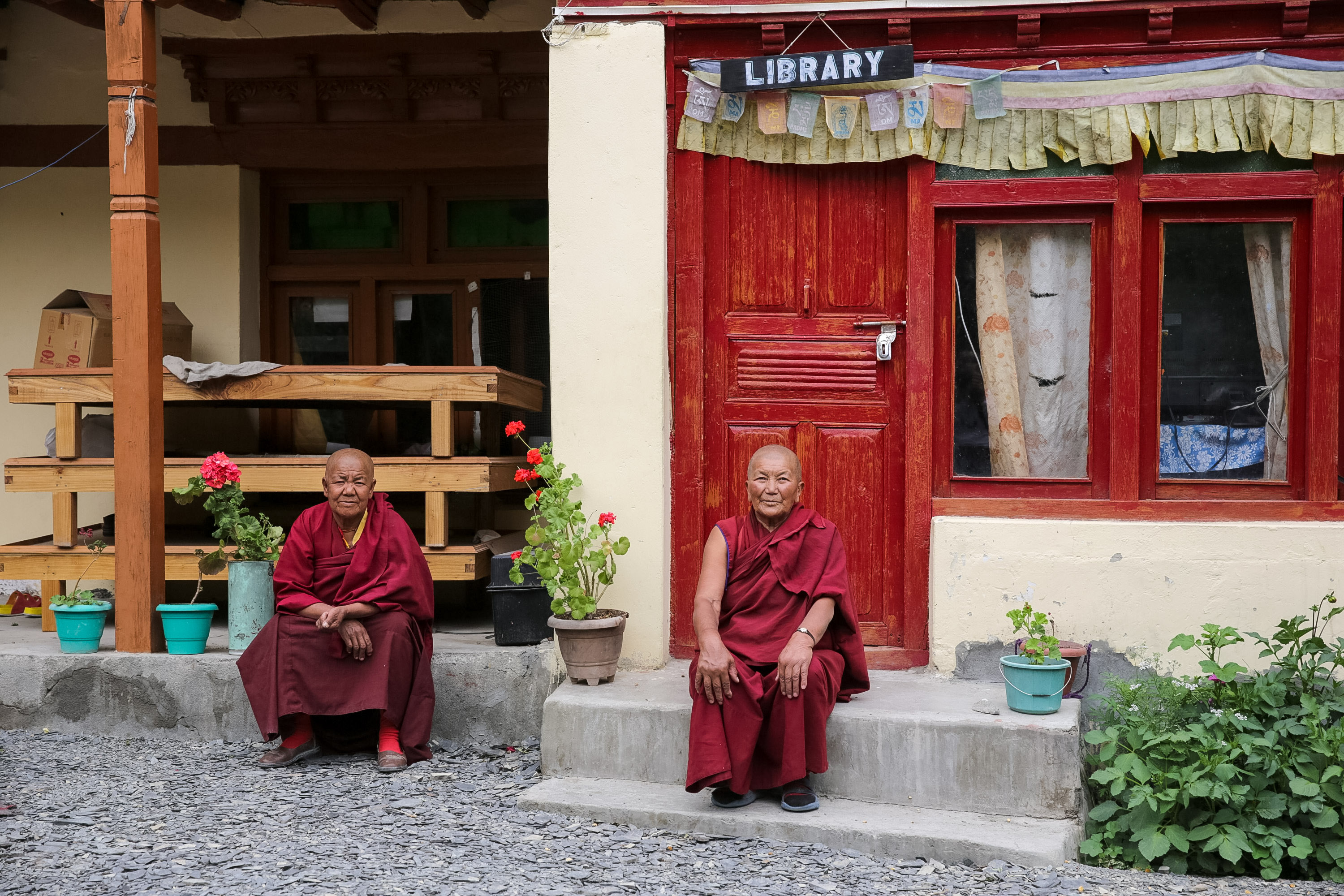 Teens look on as monks in maroon robes sit outside a red-painted building marked "Library"