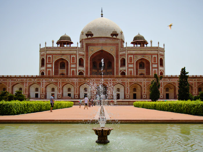 A fountain spraying in a long water channel in front of a domed sandstone tomb under a clear sky.