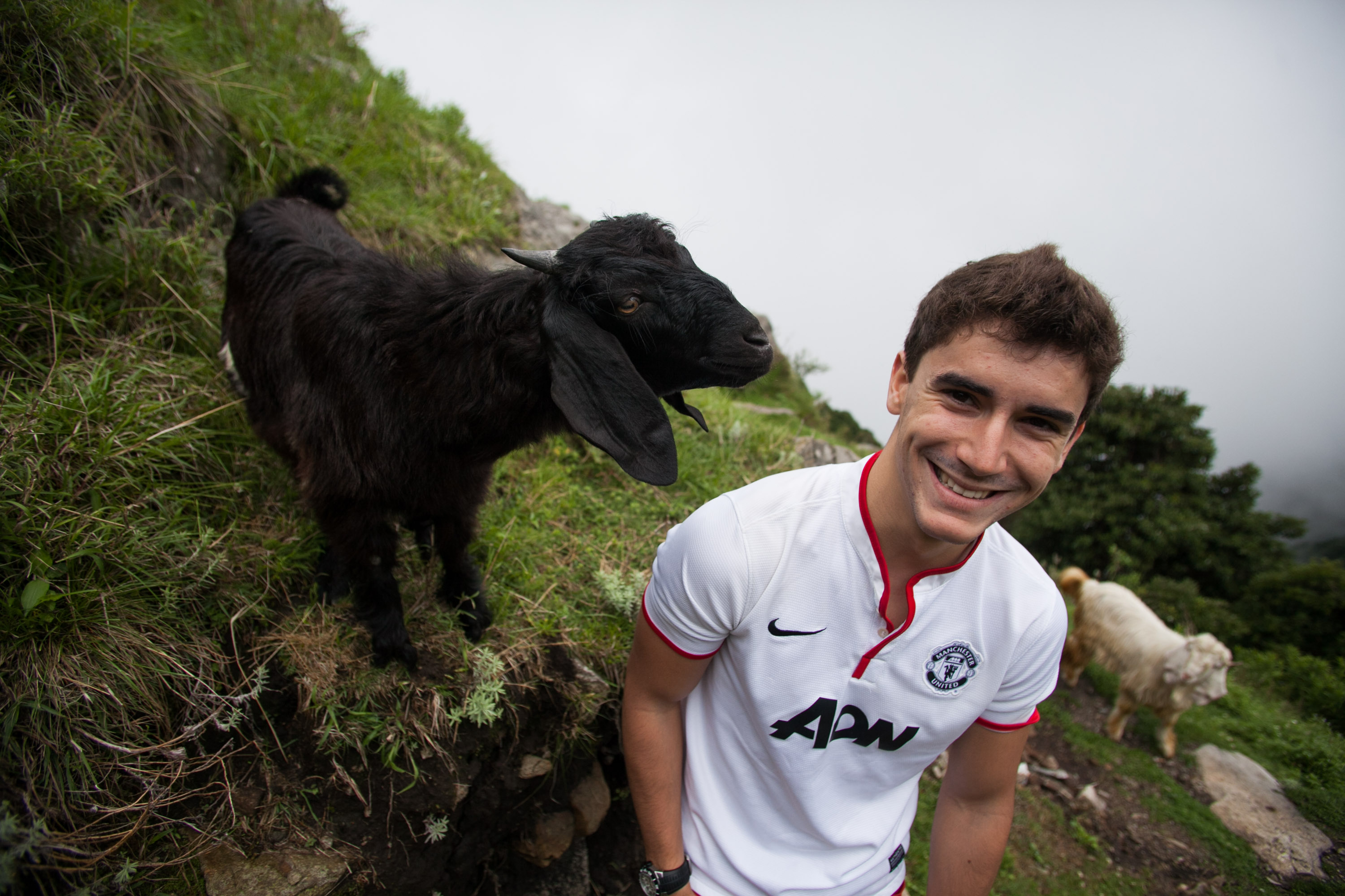 On a school trip, a student smiles on a grassy hillside next to a black goat