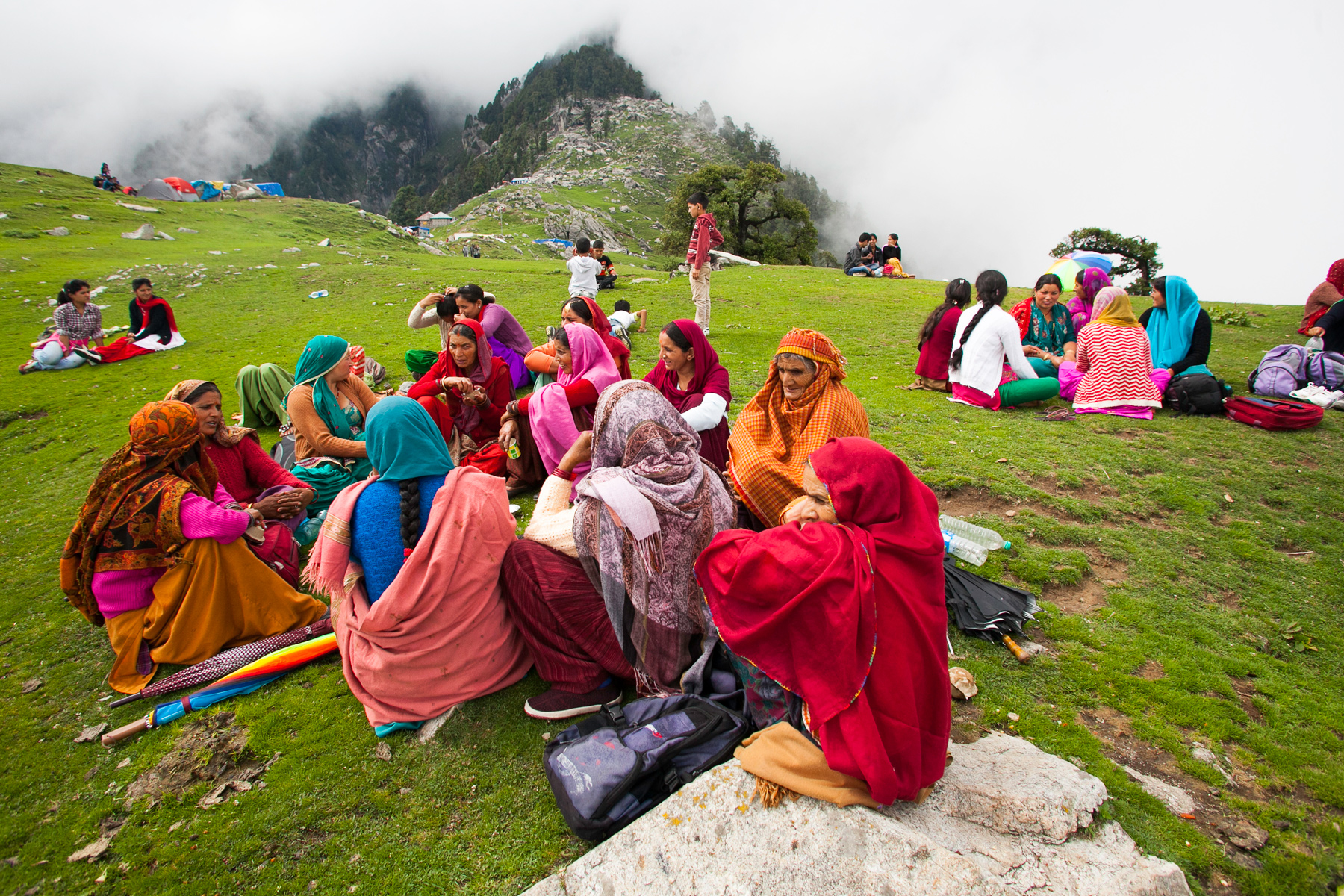 Local women in colorful traditional dress sit together on a grassy Himalayan hillside