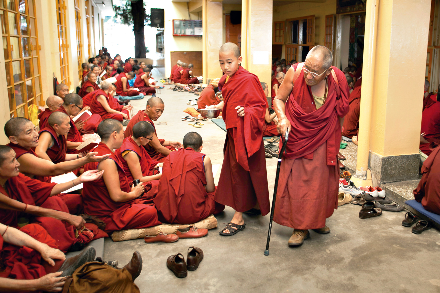 Monks in red robes sit along a corridor sharing food while an elder monk walks with a cane