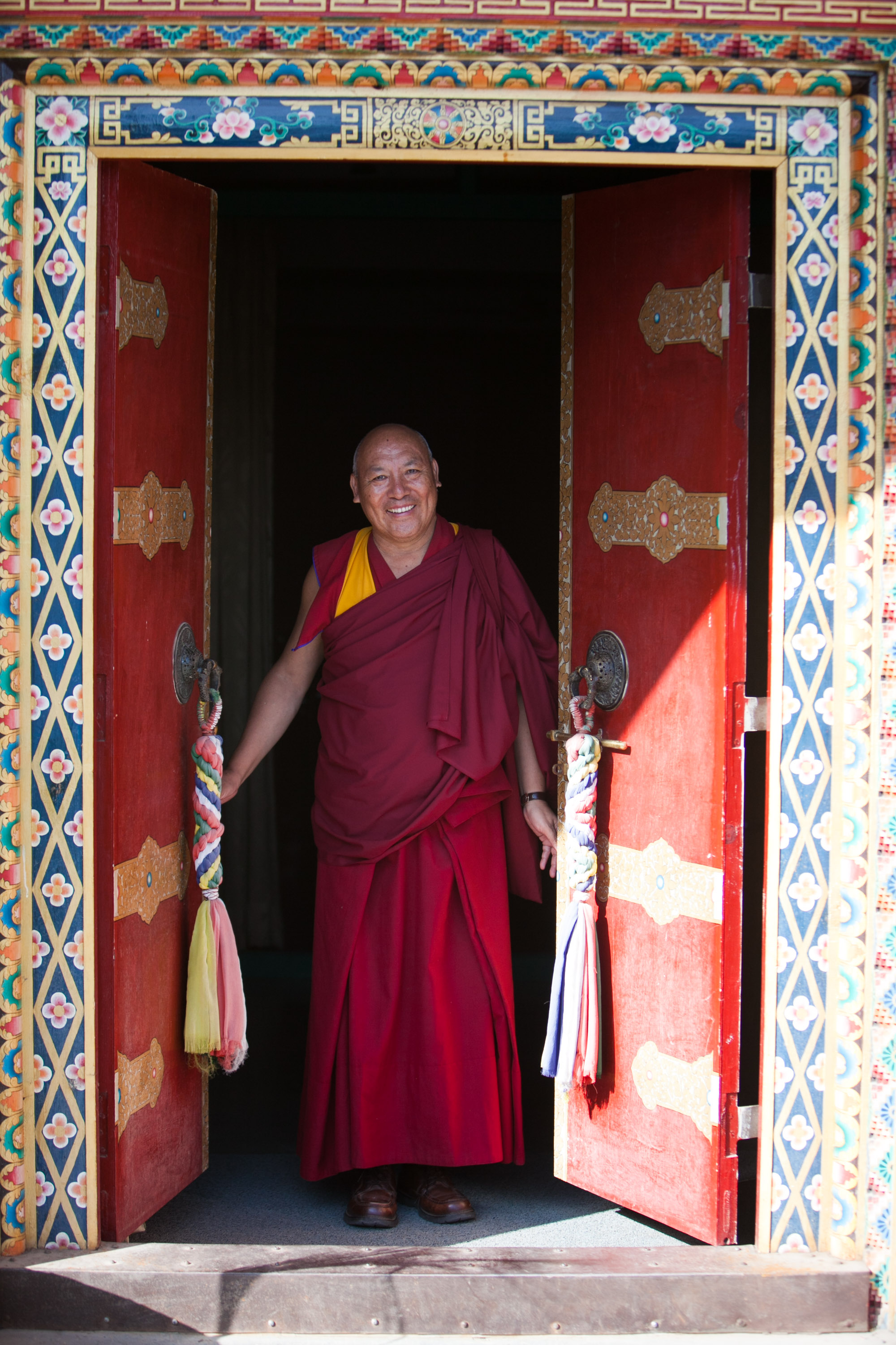 Students pause as a monk in maroon robes stands smiling in an ornately painted temple doorway