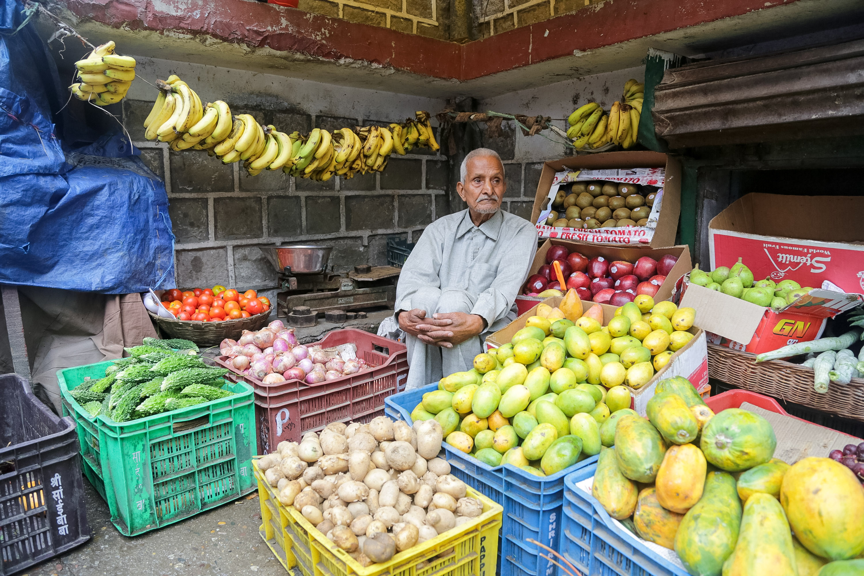 A vendor sits among crates of mangoes, papayas, potatoes and bananas at a fruit stall
