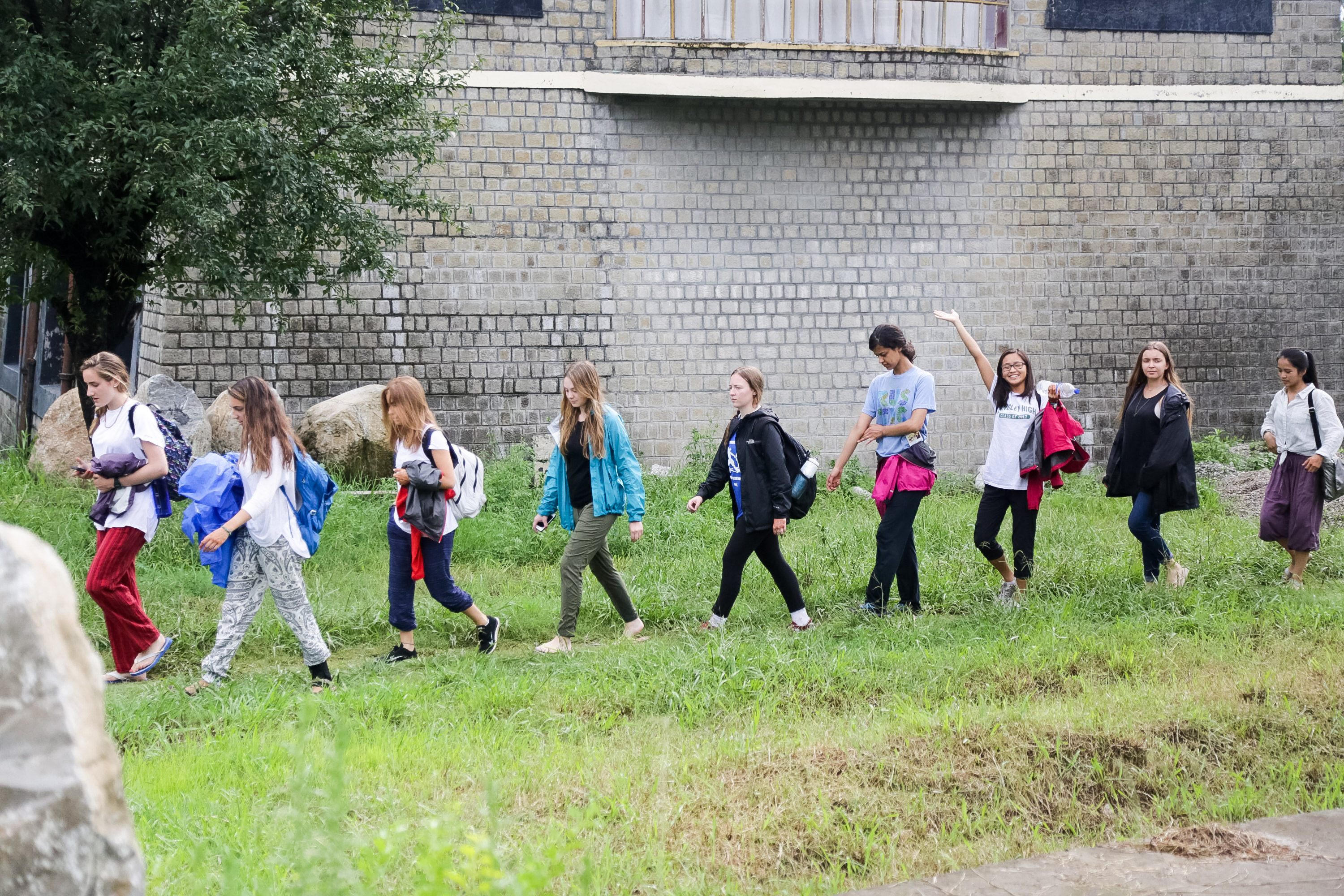Students carrying backpacks walk along a grassy path beside a stone wall