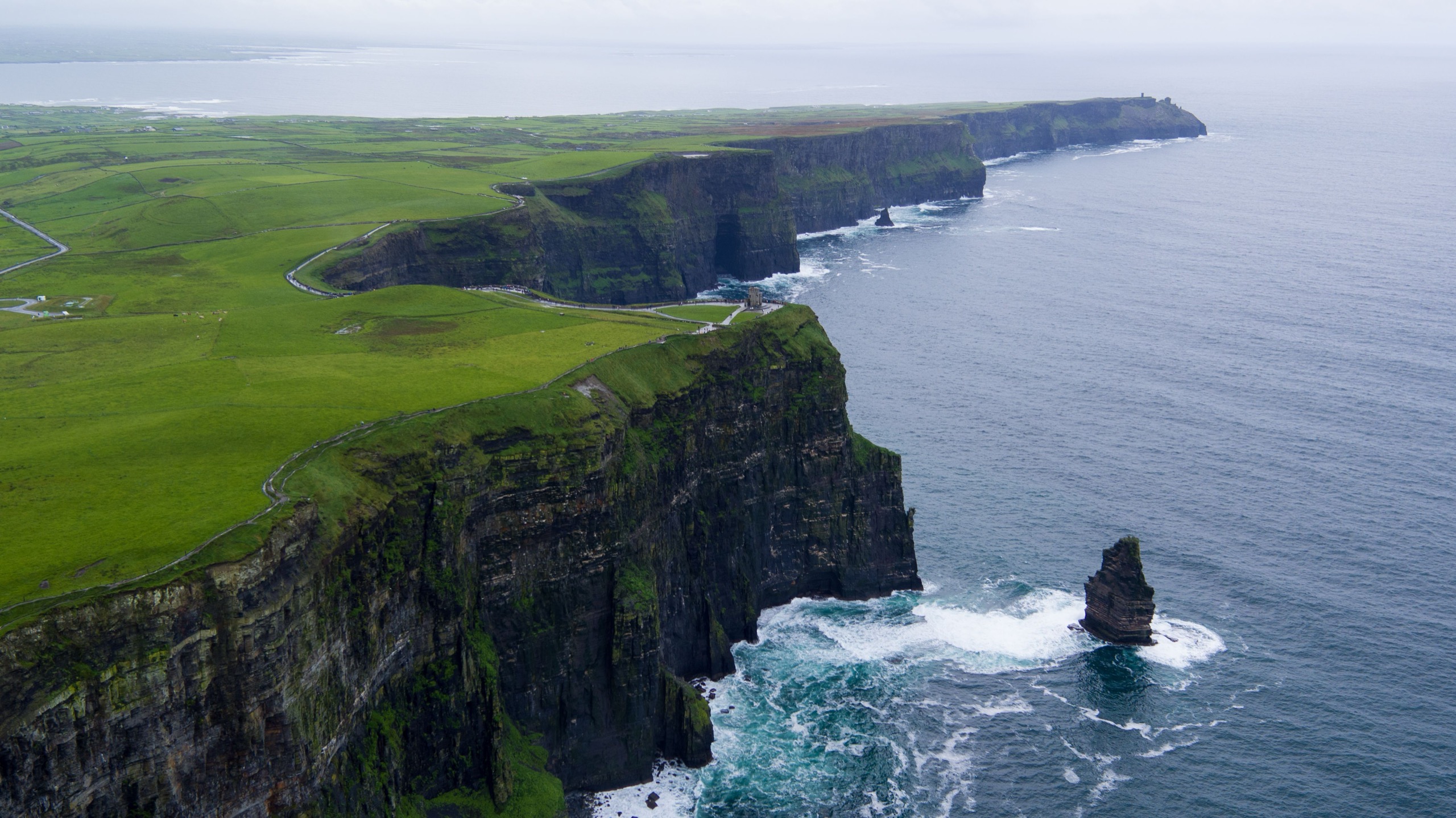 The Cliffs of Moher in County Clare, Ireland, seen from above, with green fields running to the cliff edge and a sea stack standing in the Atlantic below.