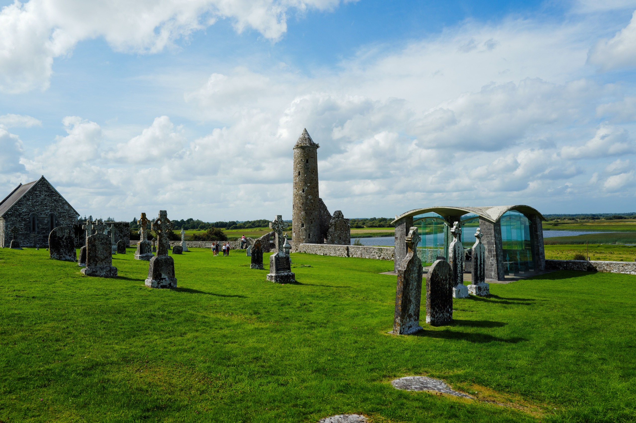 A round tower and high crosses stand among old gravestones on the green at Clonmacnoise, with the Shannon flat and silver behind them.
