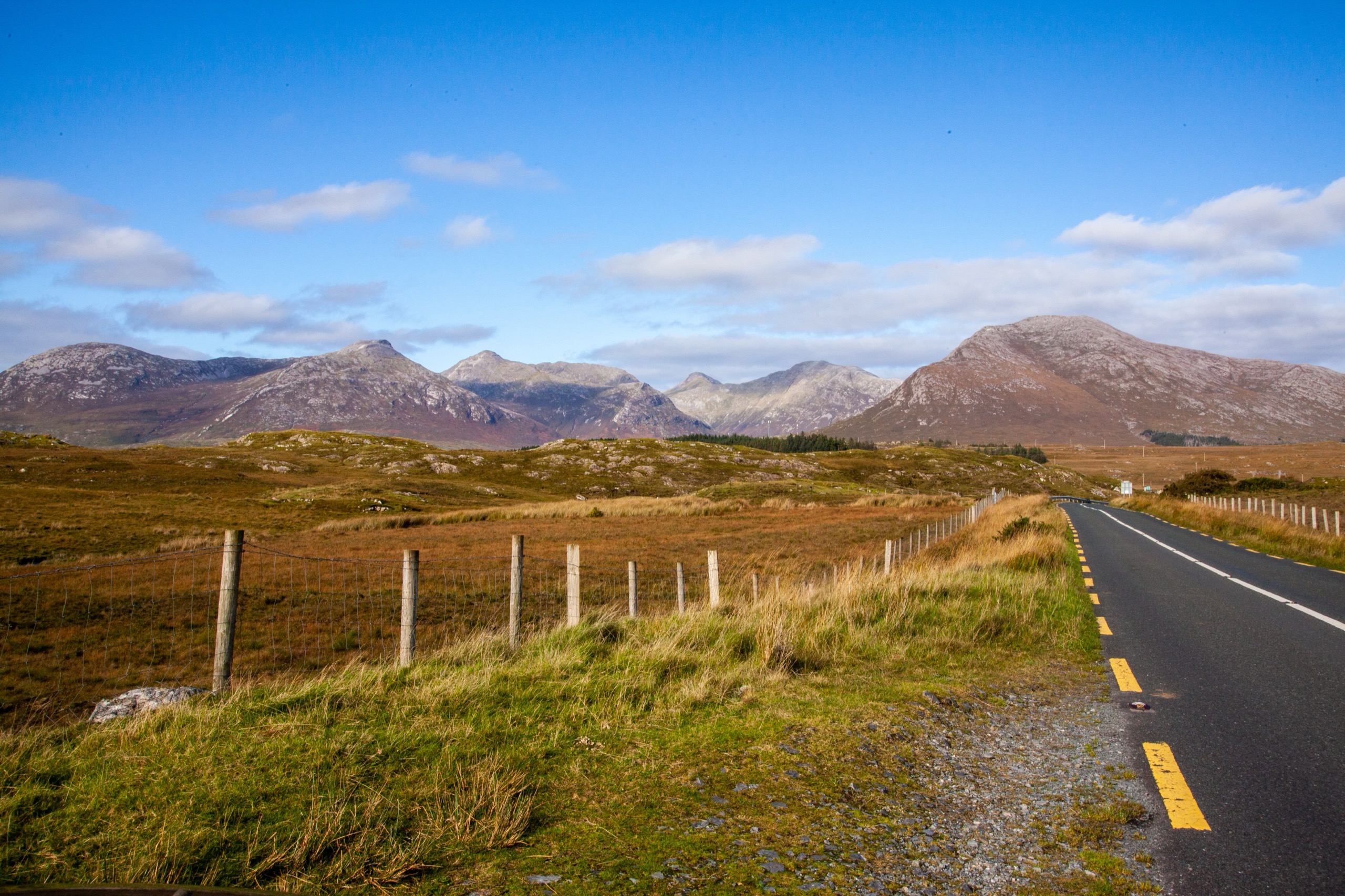 A single road runs across open bogland toward the bare rounded peaks of the Twelve Bens under a blue sky.