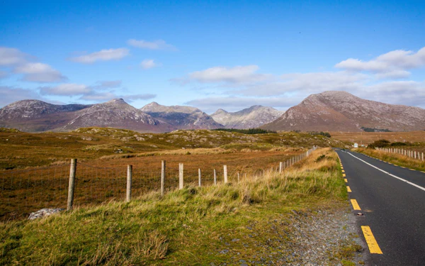 A single road runs across open bogland toward the bare rounded peaks of the Twelve Bens under a blue sky.