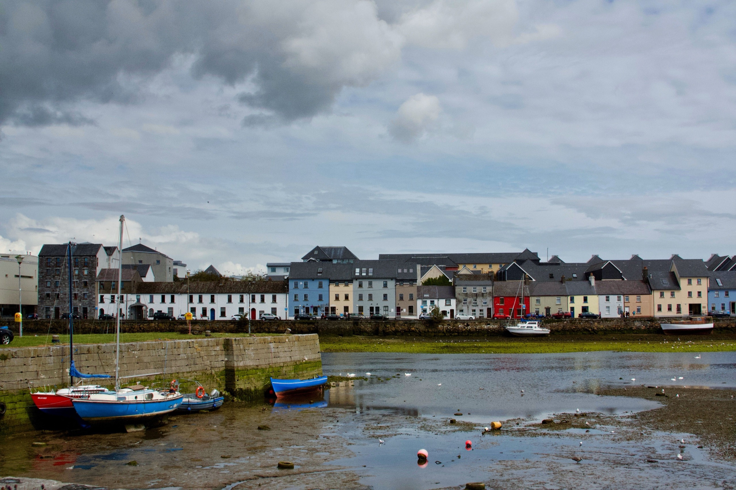 A row of painted houses along Galway's Long Walk faces the harbor at low tide, small boats resting on the mud under a heavy grey sky.