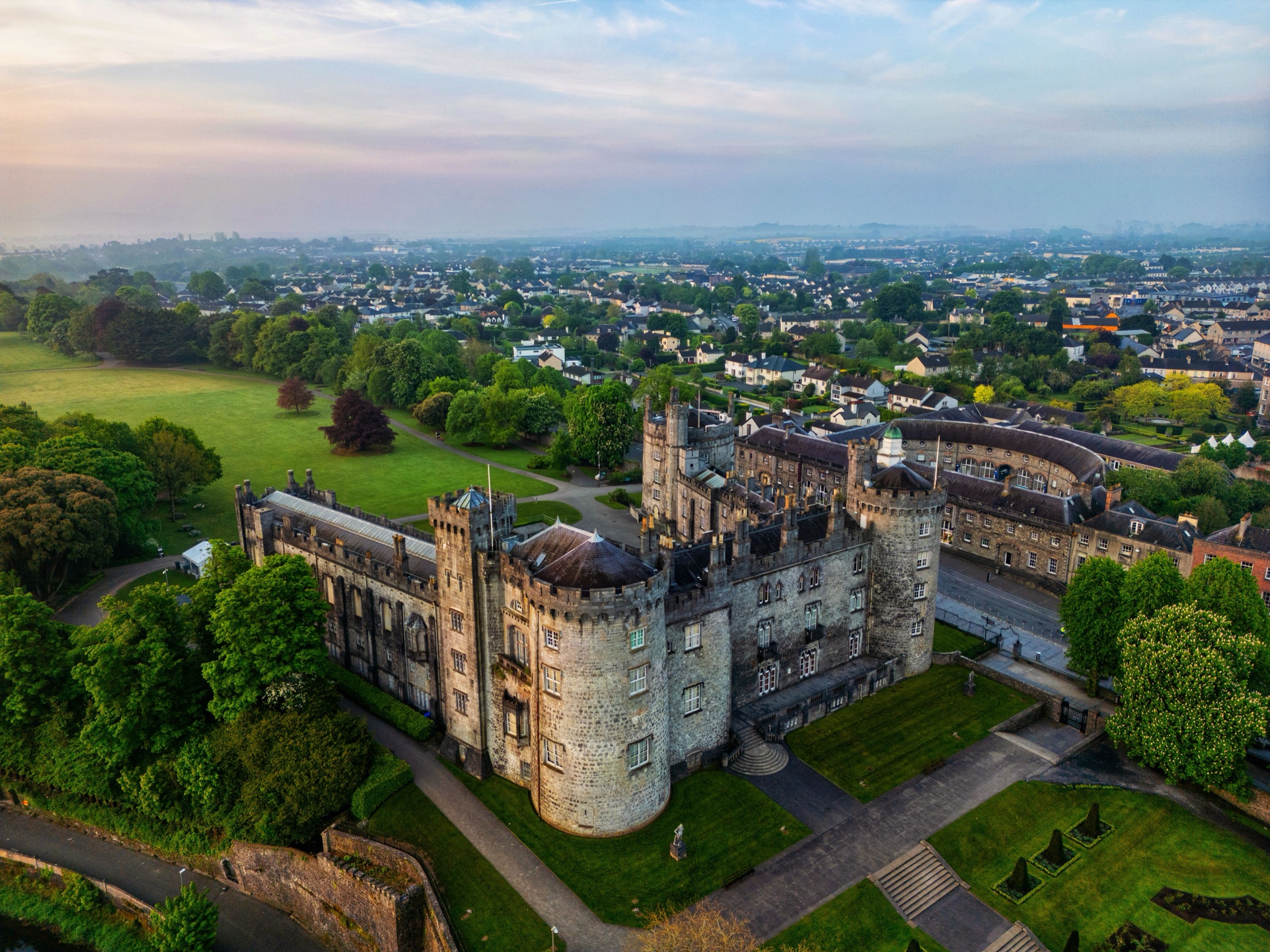 Kilkenny Castle's round towers and grey battlements rise from lawns and trees, with the town's rooftops spreading to a pale dawn horizon.