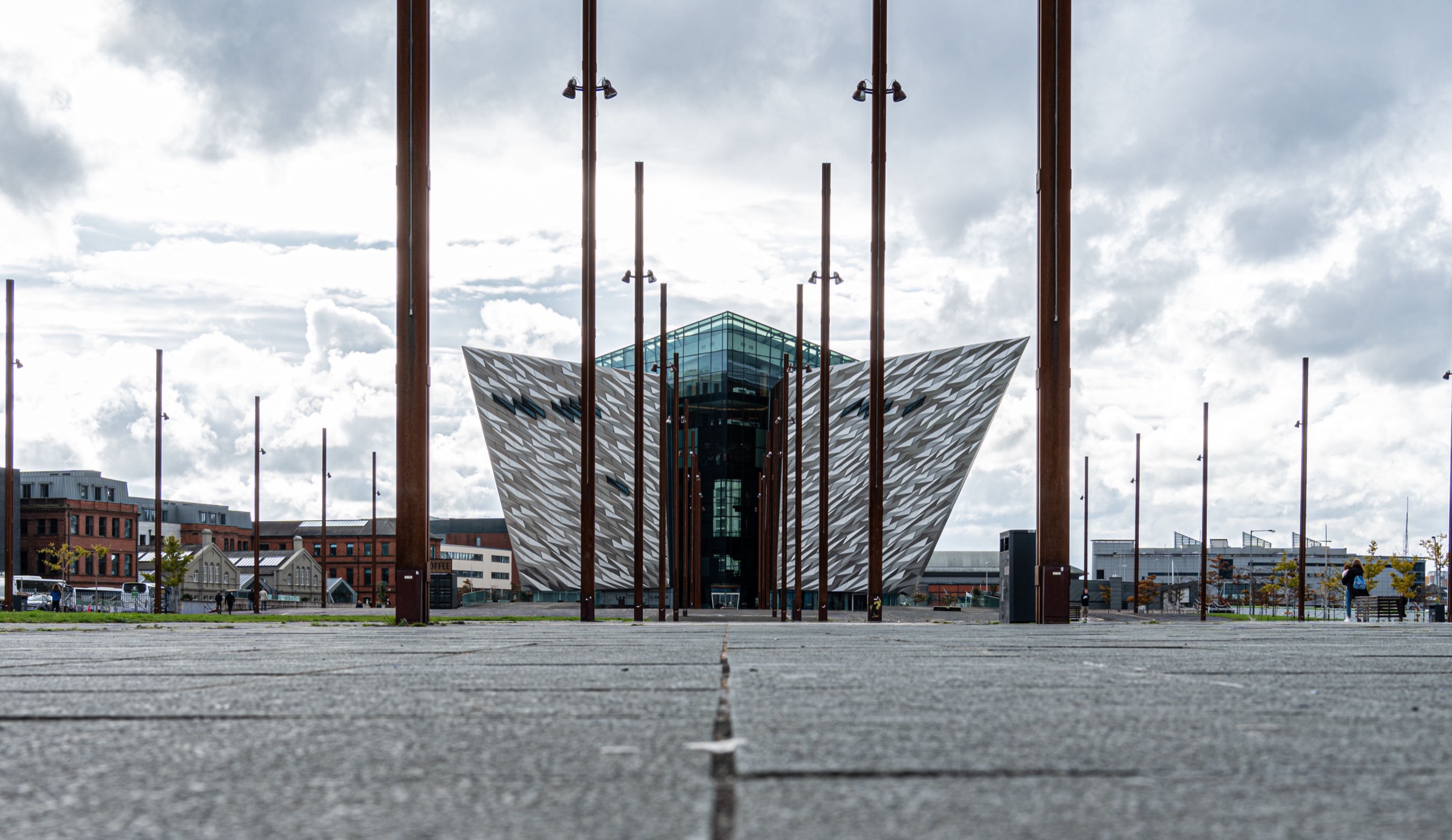 The angular silver hull-shaped building of Titanic Belfast rises behind a plaza of tall slender masts under a cloudy sky.