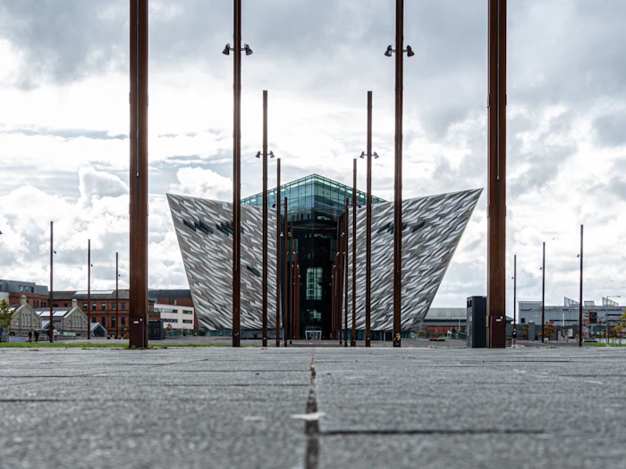 The angular silver hull-shaped building of Titanic Belfast rises behind a plaza of tall slender masts under a cloudy sky.
