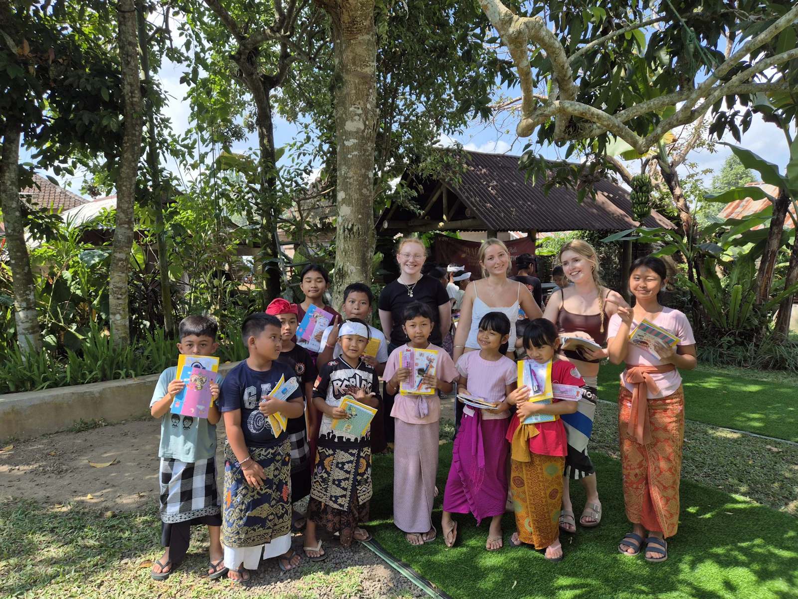 A group of teen travelers stand beside younger children in bright sarongs in a leafy garden in Tabanan, Bali