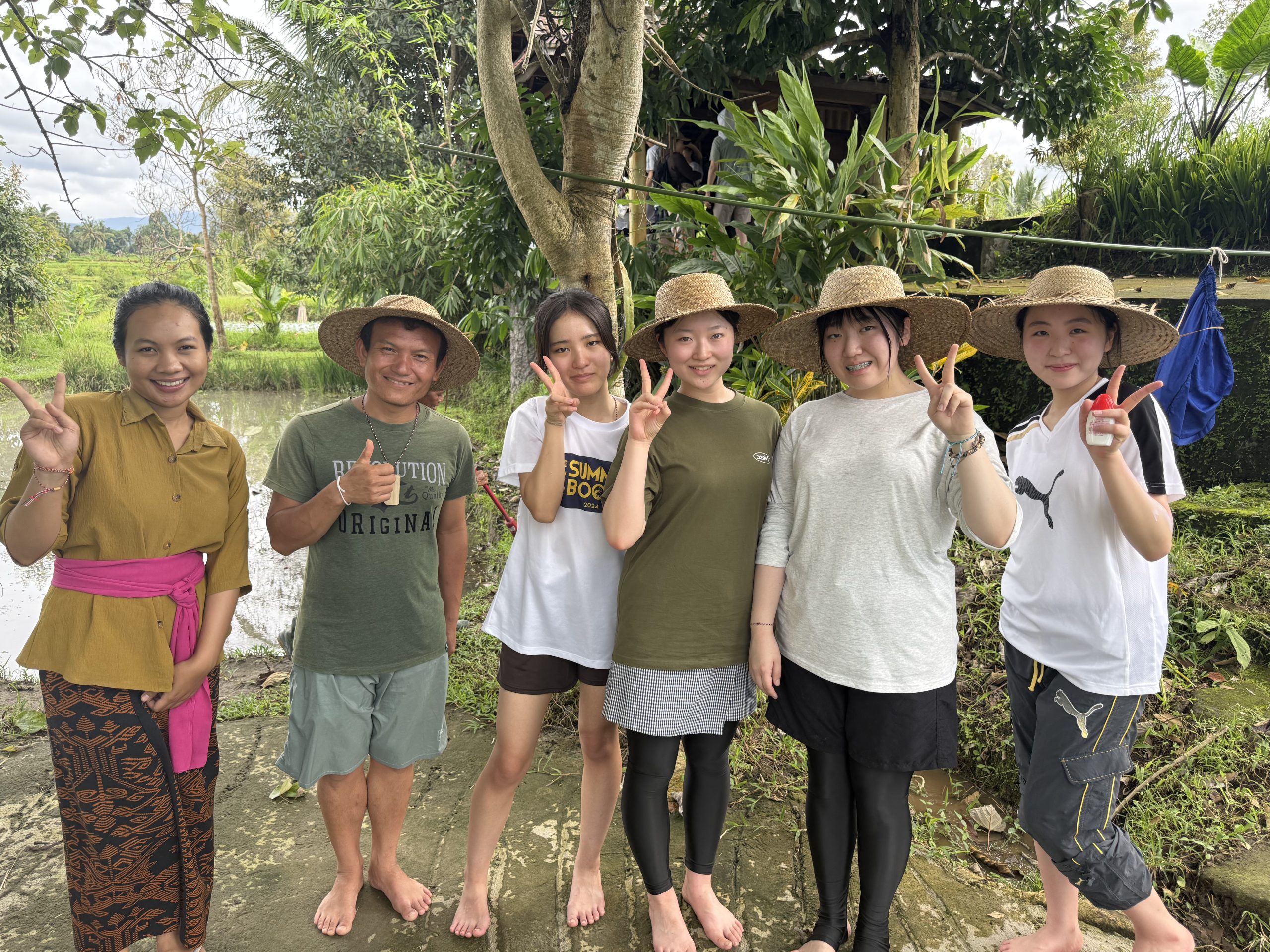 Students and local hosts in straw hats pose with peace signs beside a flooded rice paddy.