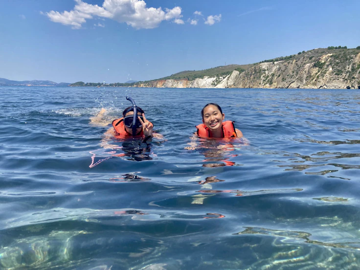 Two teen travelers in orange life vests snorkel in clear blue sea below a white cliff