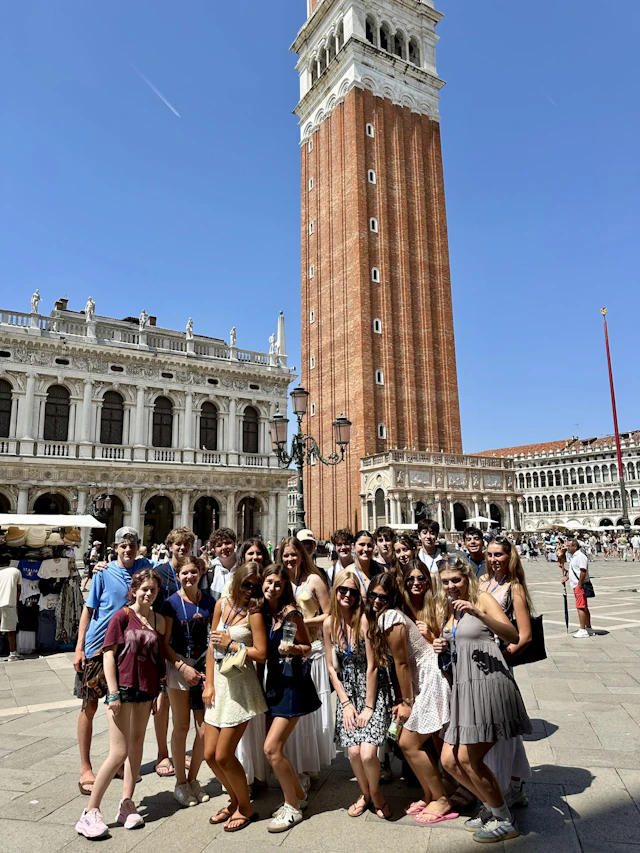 A group of teen travelers gathers in St. Mark's Square in Venice, Italy, below the tall brick bell tower on a sunny day