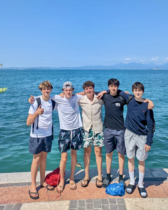 Five teen travelers stand arm in arm on a waterfront promenade on Lake Garda in Italy, with blue water and mountains behind them