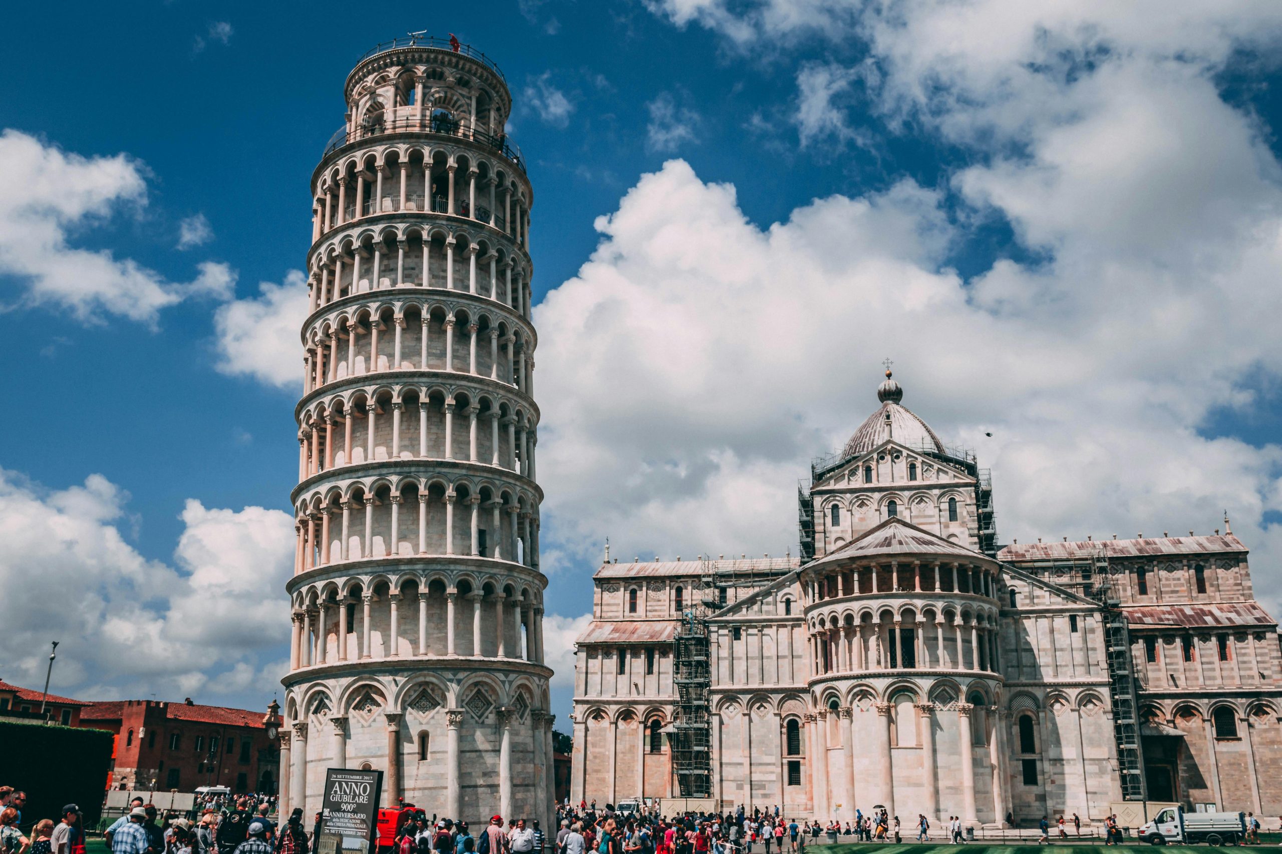 Students gather near the Leaning Tower of Pisa as it rises beside the cathedral under a partly cloudy sky