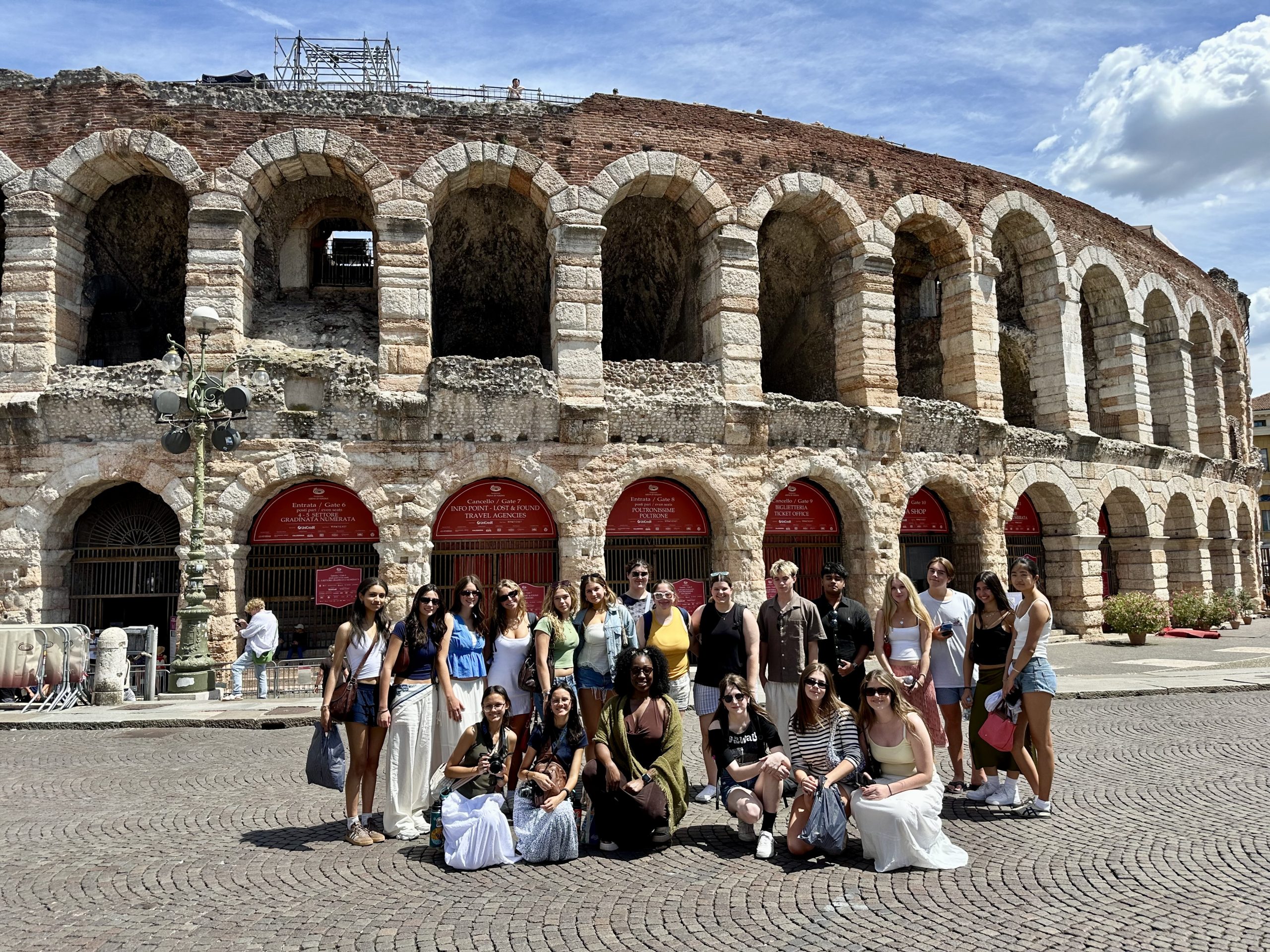 A group of teen travelers stands and kneels on the cobbles in front of the stone arches of the Roman arena in Verona