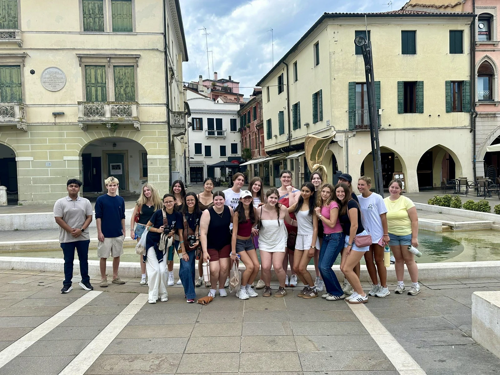 Around twenty teen travelers line up beside a low stone fountain in an arcaded town square in Italy